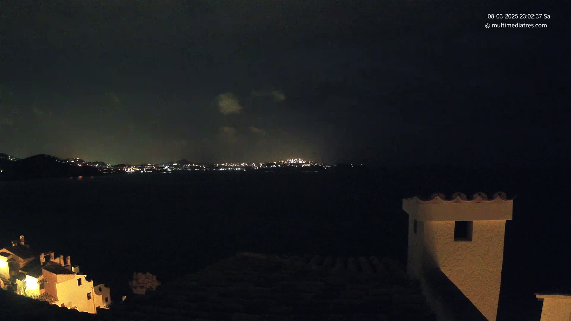 A nighttime view of a city across a dark body of water, partially obscured by clouds, with a building and chimney in the foreground.