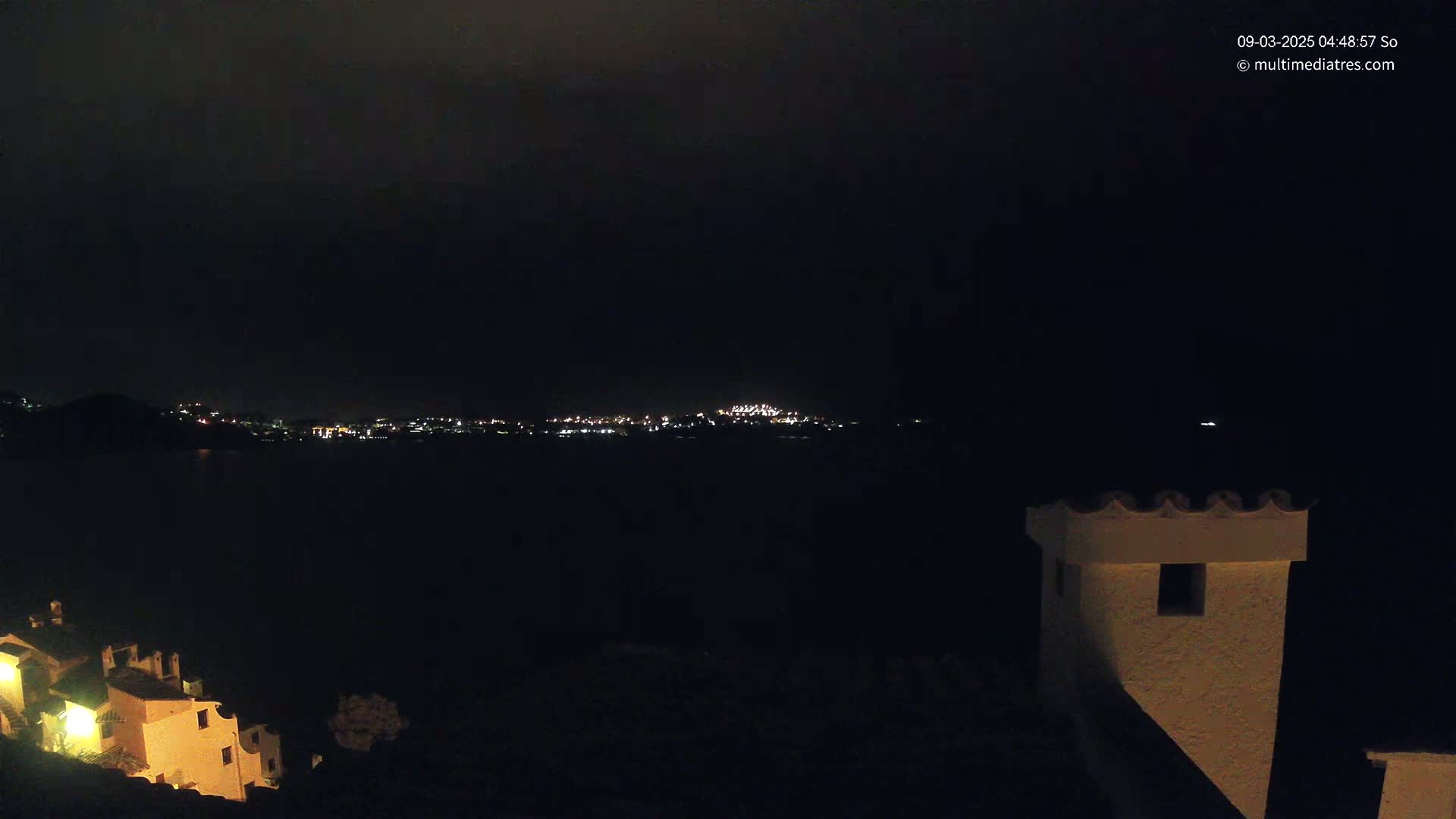 A nighttime view of a city skyline across a dark body of water, partially obscured by a dark, possibly cloudy sky, with a portion of a building in the foreground.