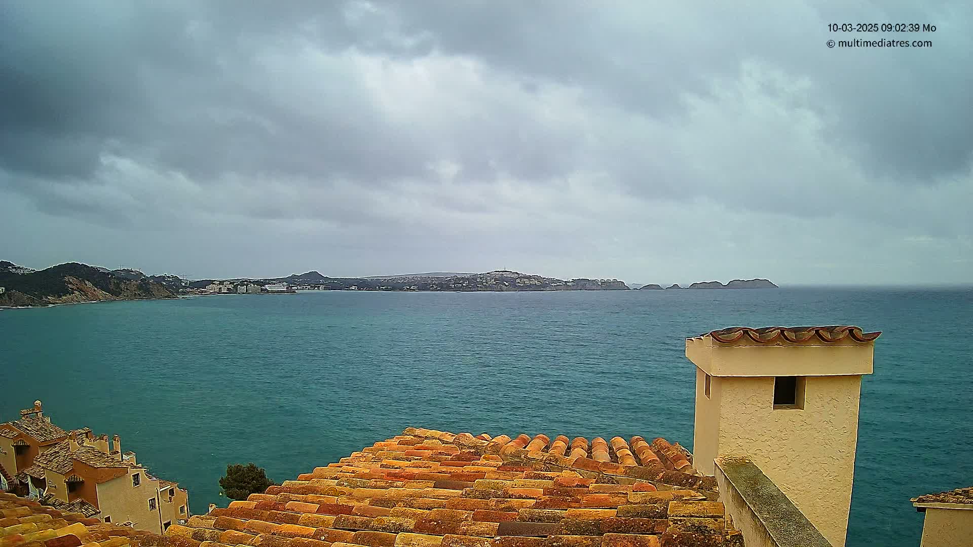 A teal-colored sea stretches to a distant coastline under a cloudy sky, viewed from a terracotta roof with a light-beige chimney.