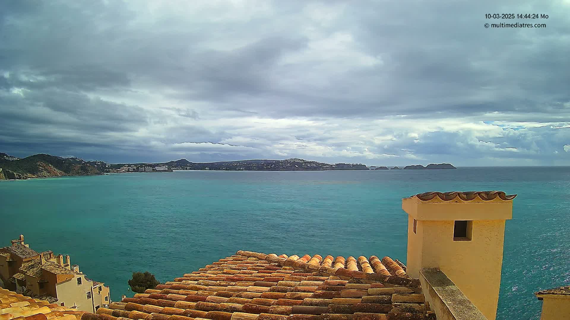 A teal-colored sea stretches to a distant coastline under a cloudy sky, viewed from a terracotta roof with a pale yellow chimney.