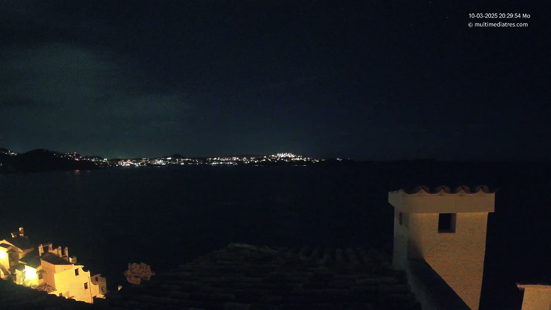 A nighttime view from a rooftop shows a dark body of water separating the viewer from a distant city illuminated with lights under a clear, dark sky.