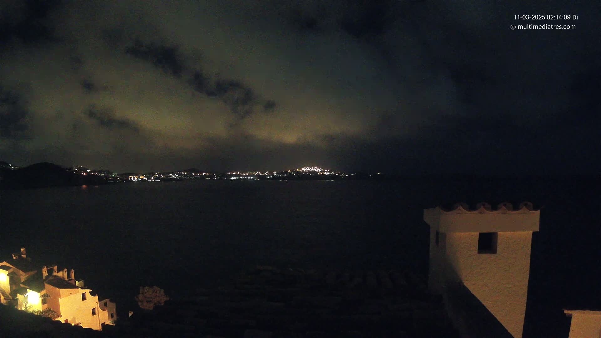 A nighttime coastal scene shows a dark body of water with a faintly lit town across it under a cloudy sky, partially illuminated buildings on the left, and a white structure on the right.