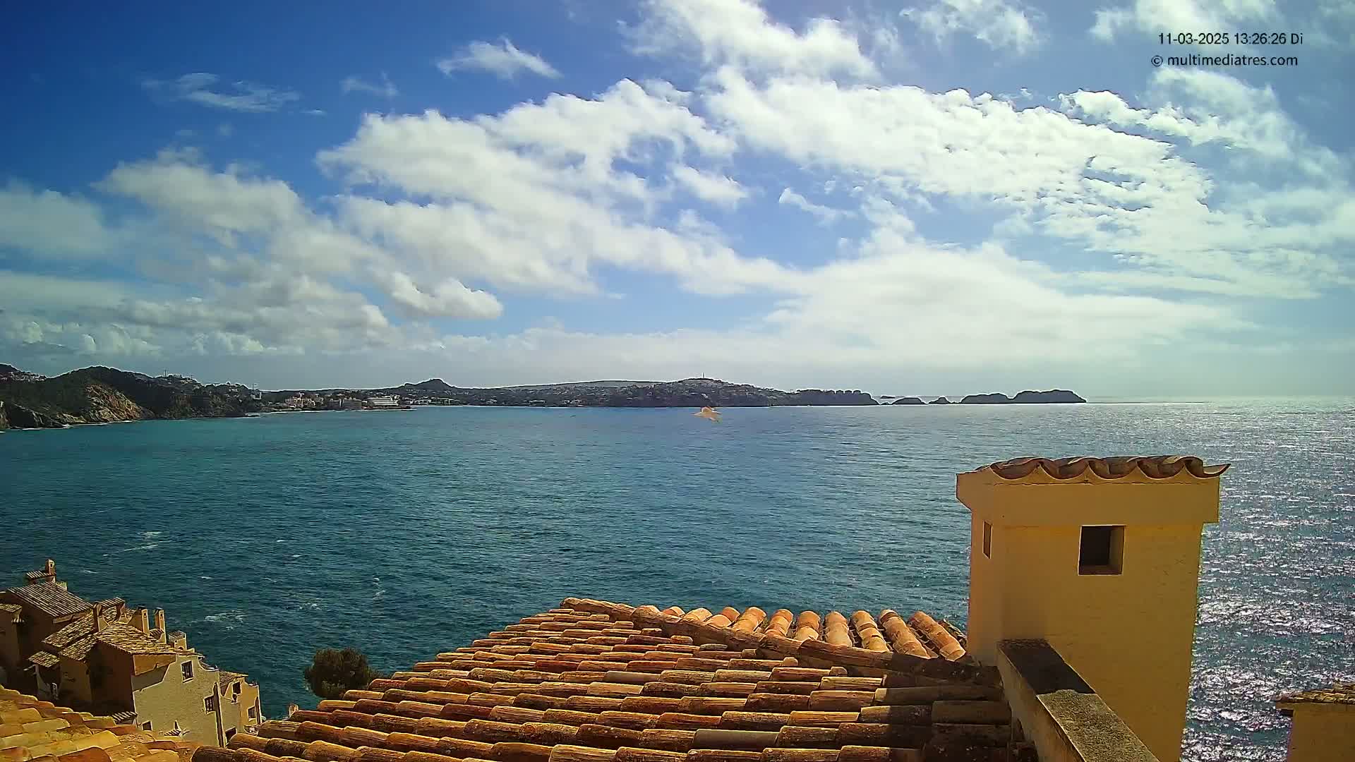 A partly cloudy sky overlooks a calm blue sea, with a terracotta-tiled roof and a small building in the foreground and a distant coastline.