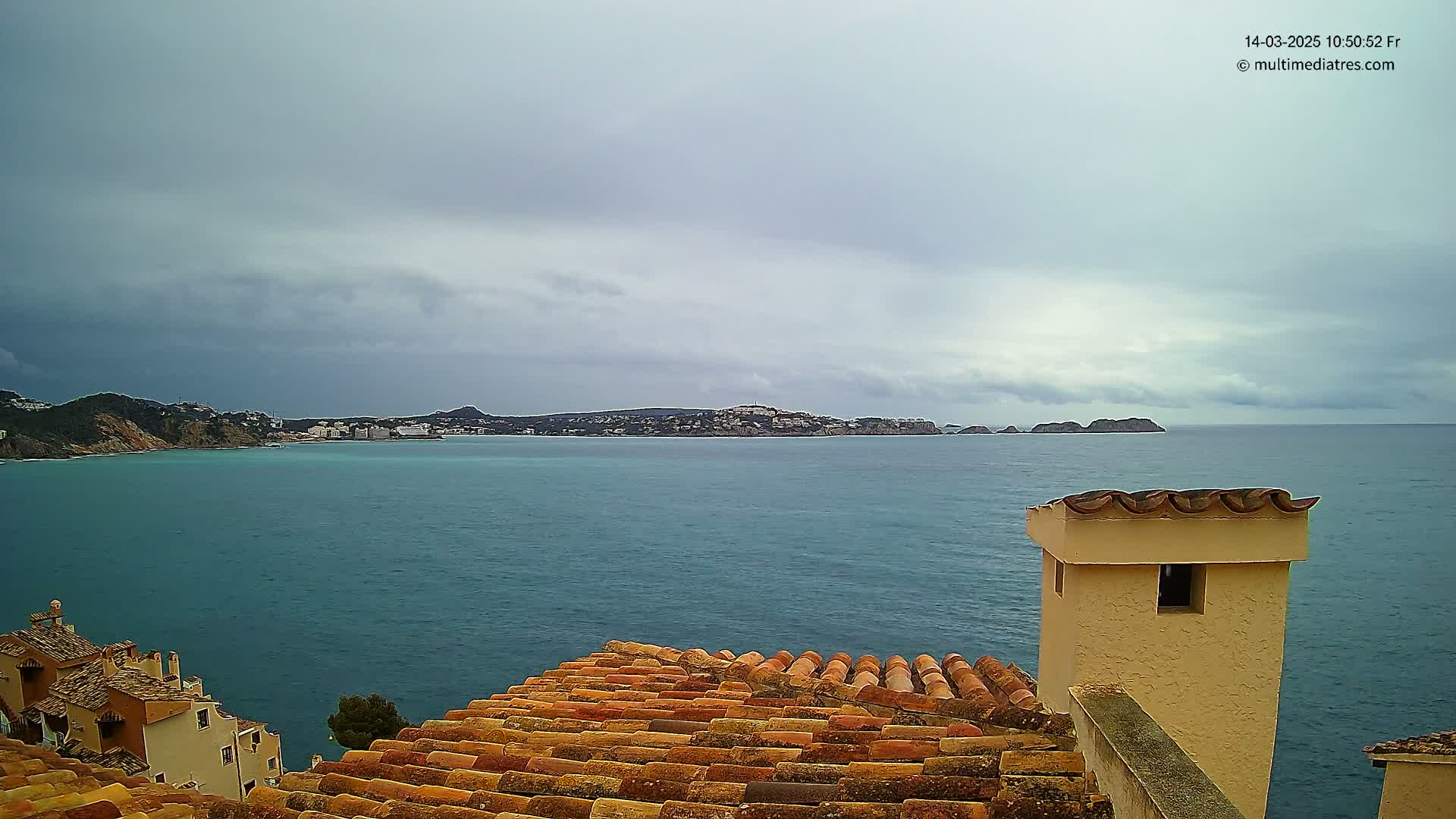 A view from a tiled roof shows a calm, overcast ocean with a distant coastline under a cloudy sky.