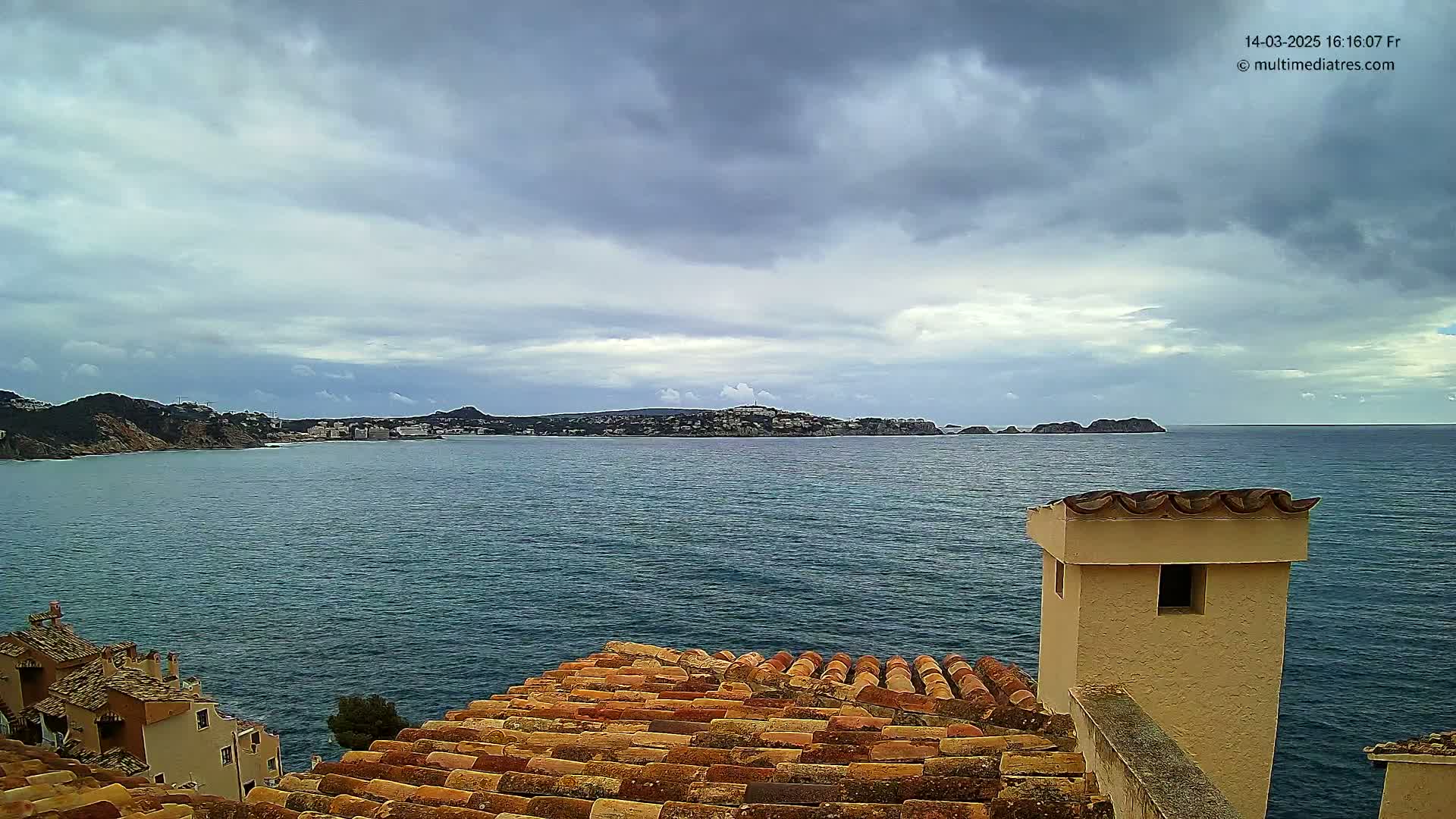 A view of a calm sea with a distant coastline under a cloudy sky, seen from a building's terracotta roof with a chimney in the foreground.