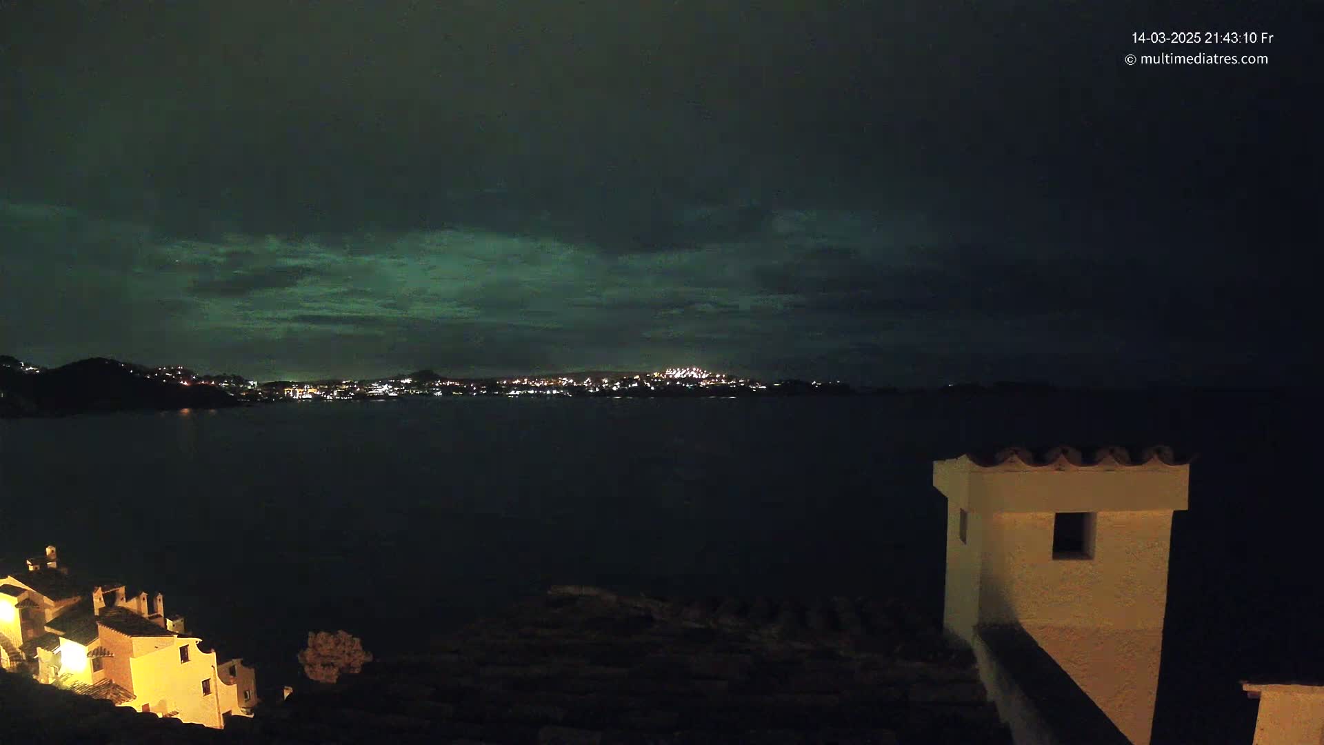 A nighttime coastal scene shows a city's lights across a dark body of water under a clouded, greenish sky.