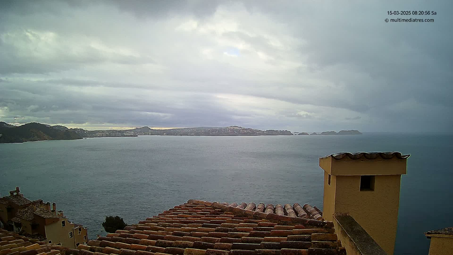 A view of a calm sea with a distant town and islands under a cloudy sky, seen from a tiled rooftop with a chimney.