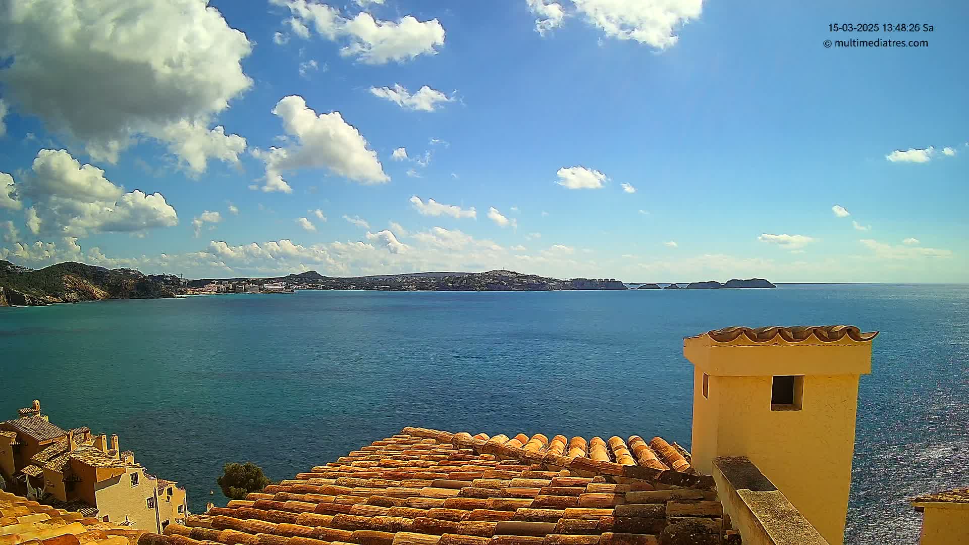 A partly cloudy blue sky overlooks a calm blue sea with a distant coastline, viewed from a tiled rooftop with a small, light-yellow chimney.