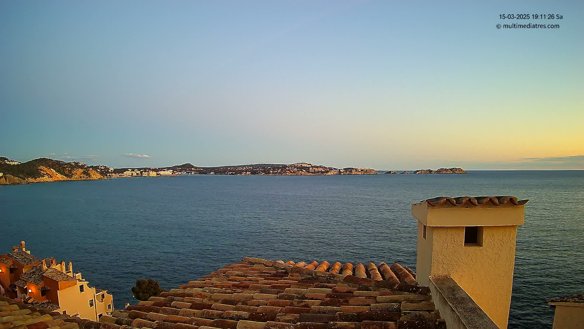 A coastal scene at sunset shows a calm sea with a distant town nestled on a peninsula, viewed from a tiled rooftop with a chimney.