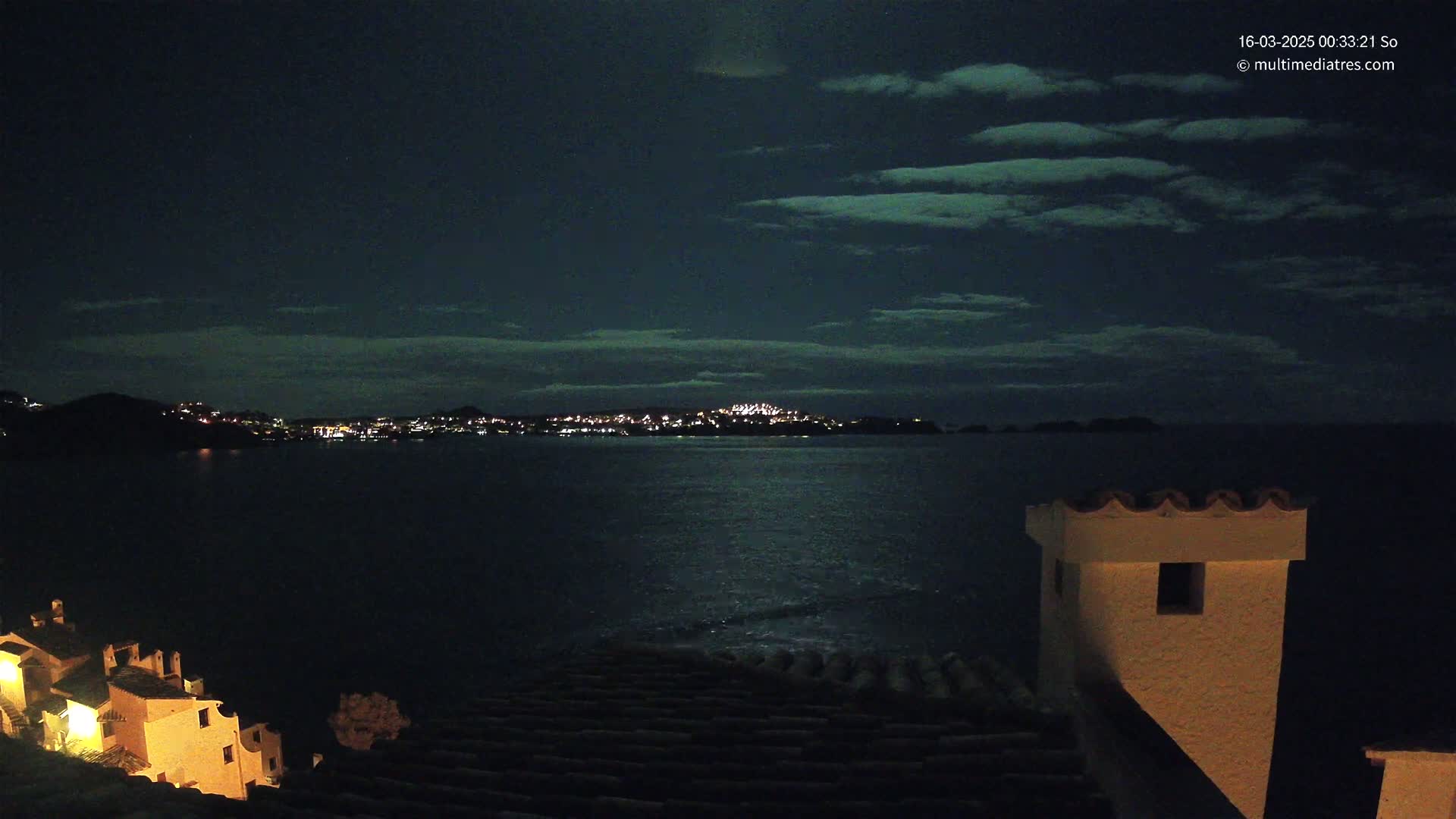 A nighttime coastal scene shows a moonlit body of water with a town’s lights visible in the distance under a partly cloudy sky.