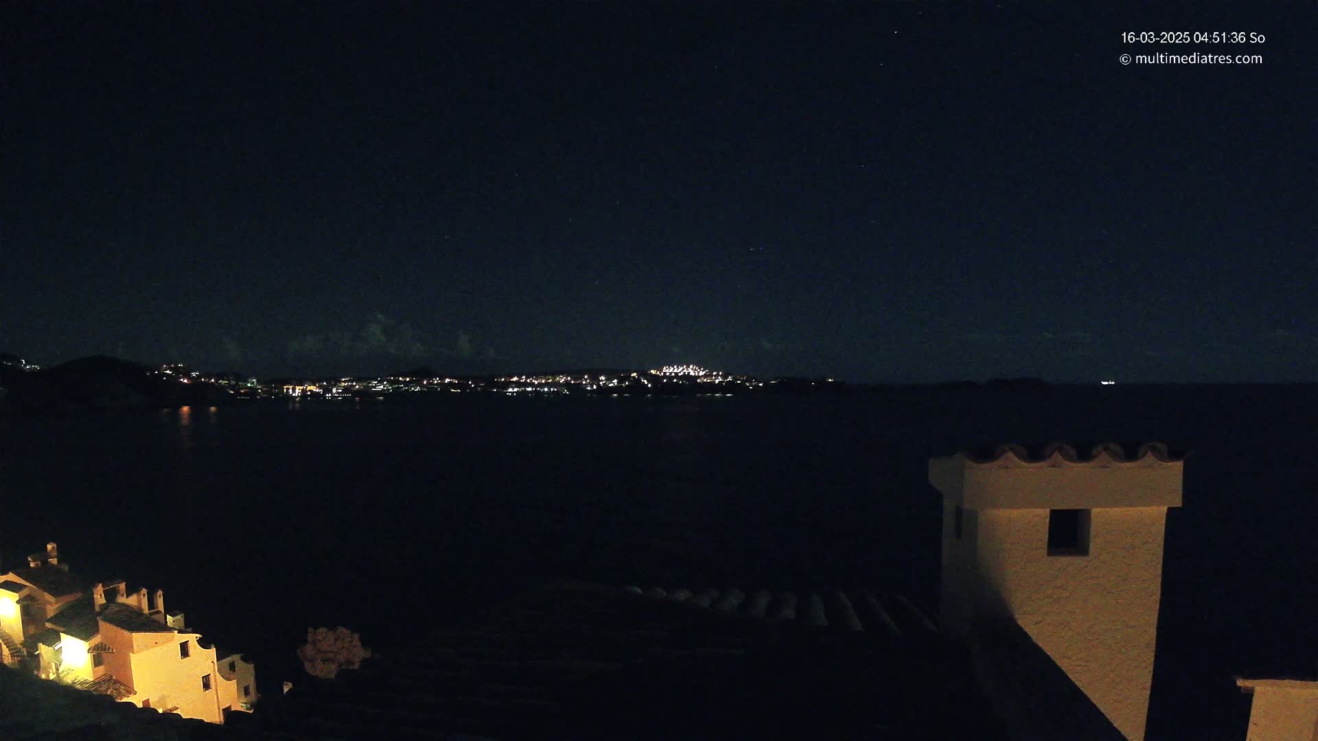 A nighttime view from a hilltop shows a dark body of water with a distant city illuminated by lights under a clear, starry sky.
