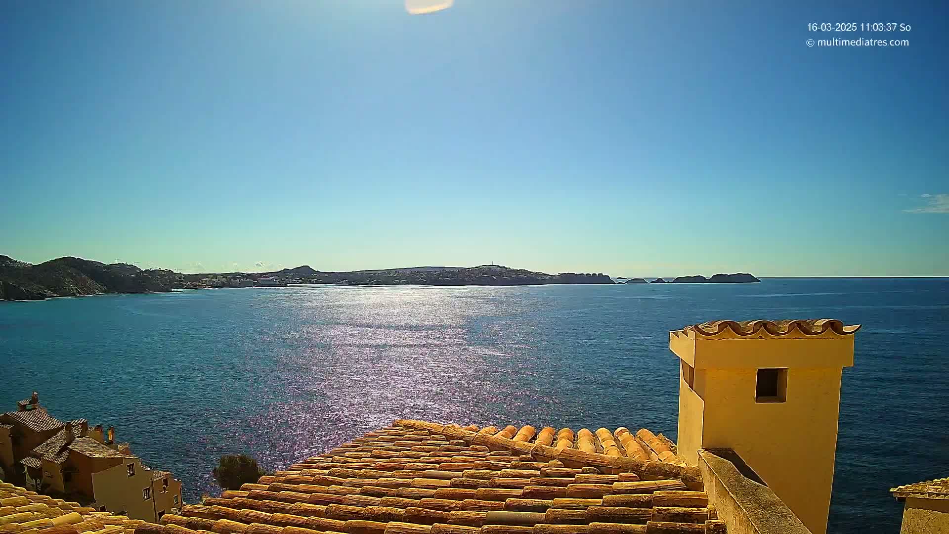 A sunlit view of a calm, blue sea from a terracotta rooftop shows distant hills and a portion of a building with a chimney.