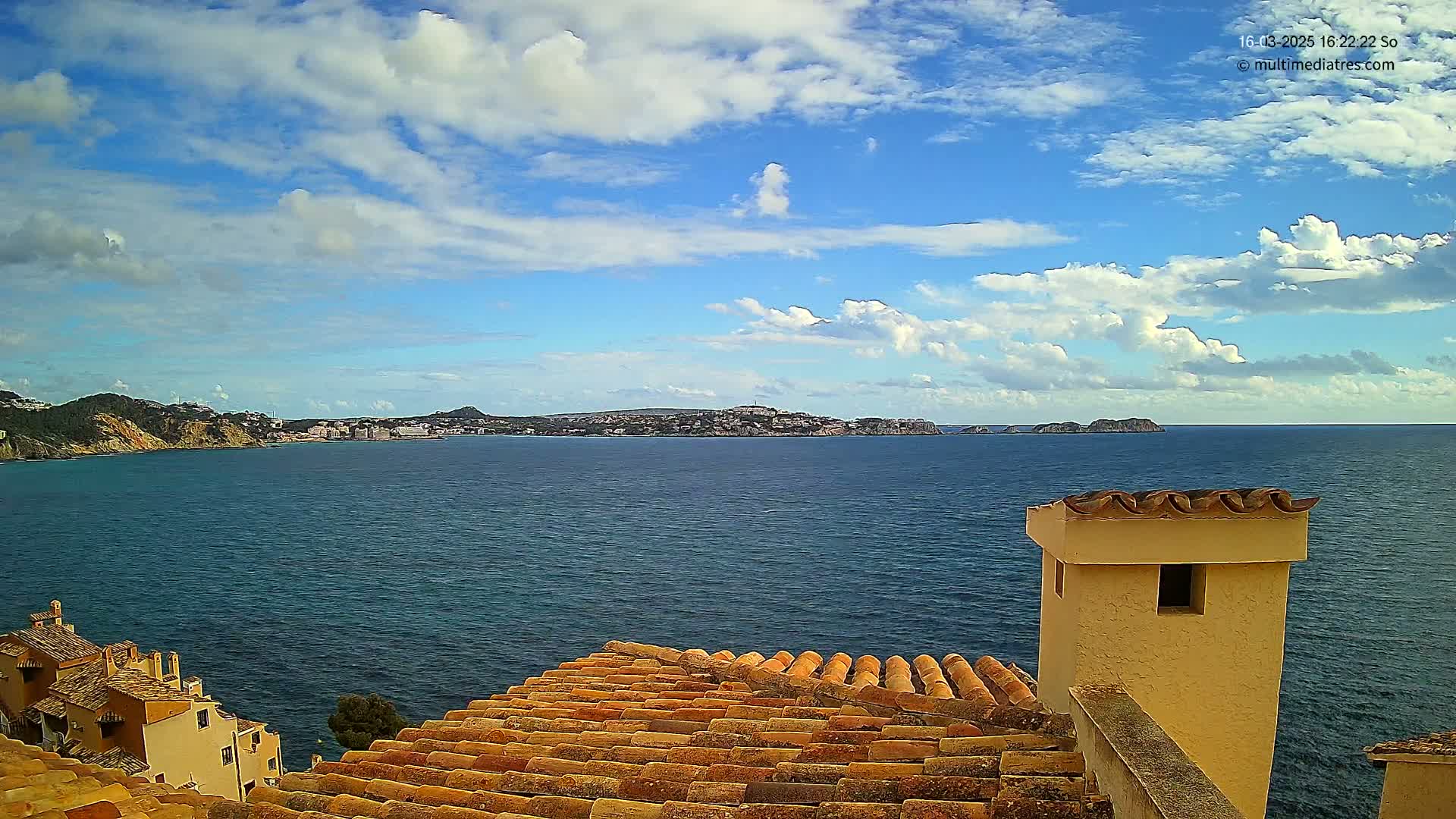 A partly cloudy sky overlooks a calm ocean, with a distant town nestled between hills and a tiled roof in the foreground.