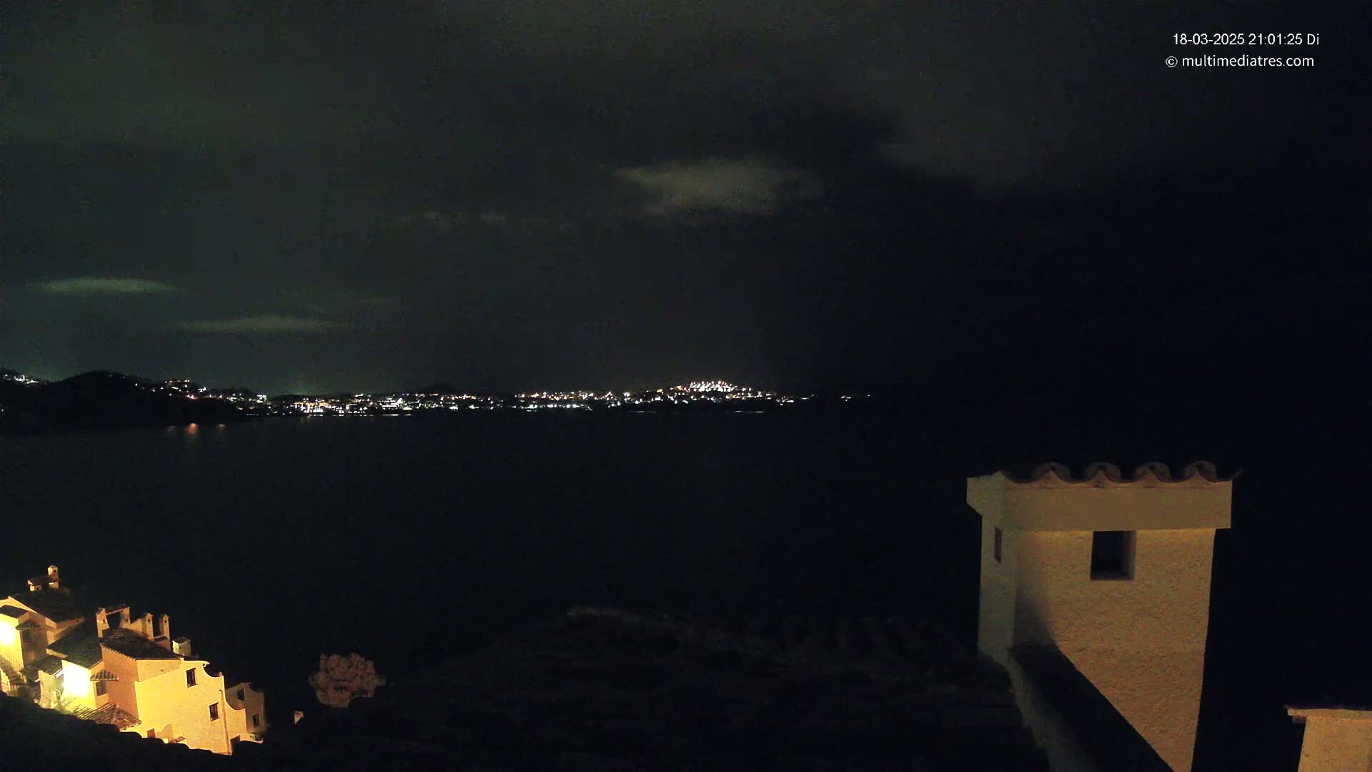 A nighttime view of a city's lights across a dark body of water, partially obscured by a cloudy sky, with a section of a lit building in the foreground.