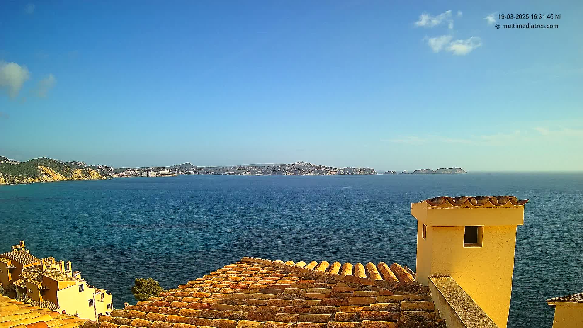 A clear blue sky overlooks a calm blue sea, a distant coastline with buildings, and a portion of a terracotta-tiled roof with a pale yellow chimney.
