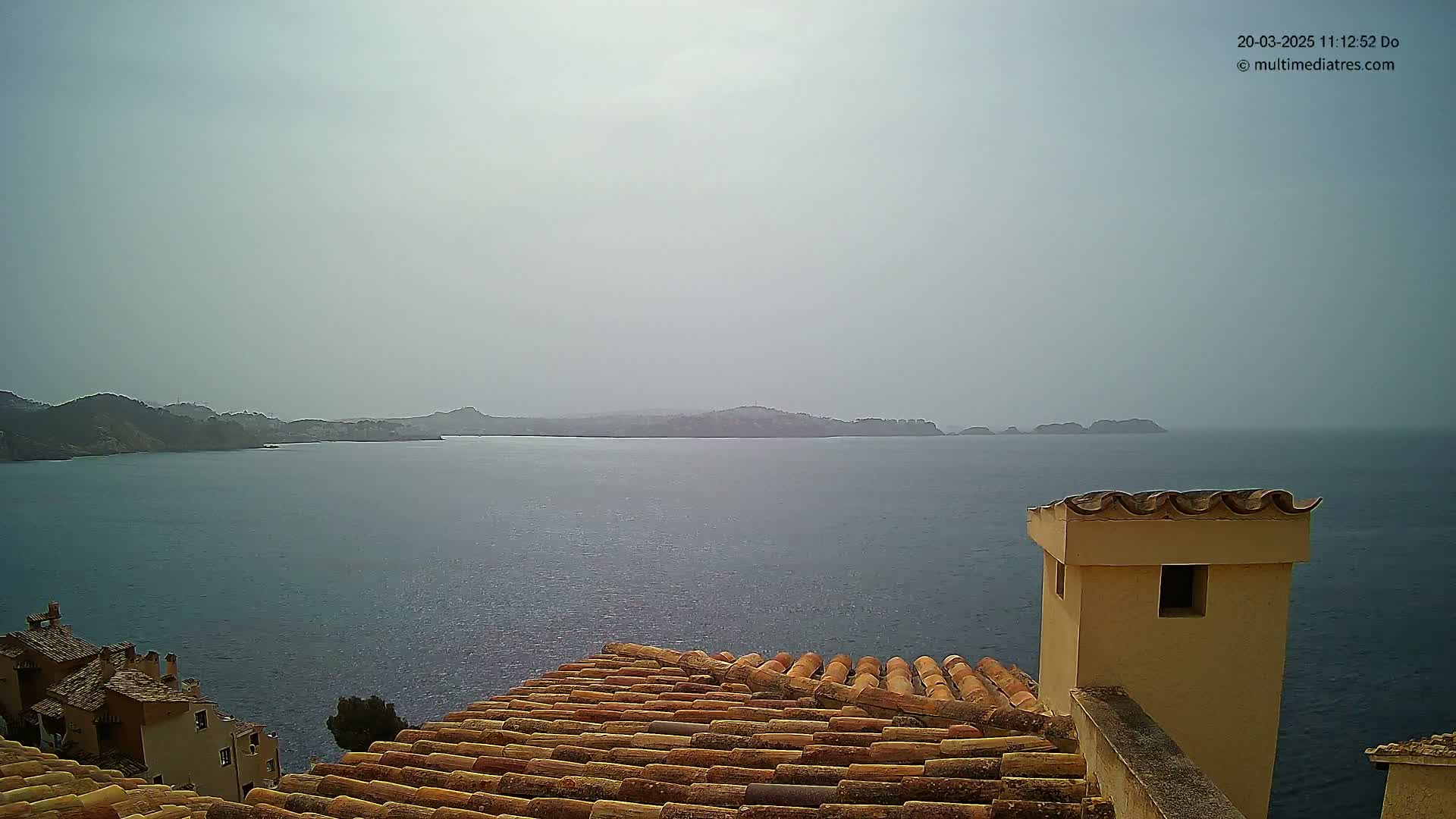 A hazy view of a calm sea and distant coastline is seen from a tiled rooftop with a small chimney.