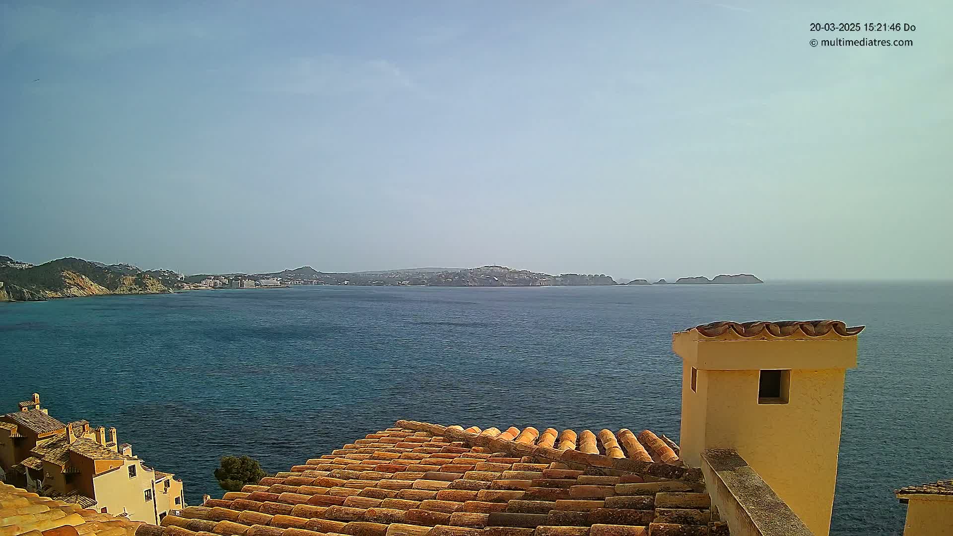 A coastal view shows a calm blue sea under a clear sky, with a terracotta roof and chimney in the foreground, and a distant town nestled into the coastline.