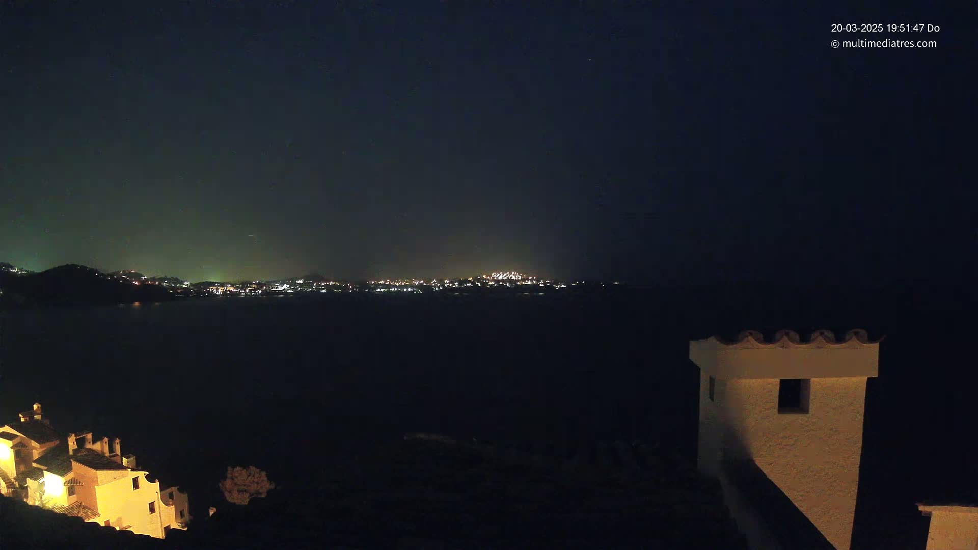 A nighttime view of a town's lights across a dark body of water, seen from a hilltop with buildings illuminated in the foreground under a clear, dark sky.
