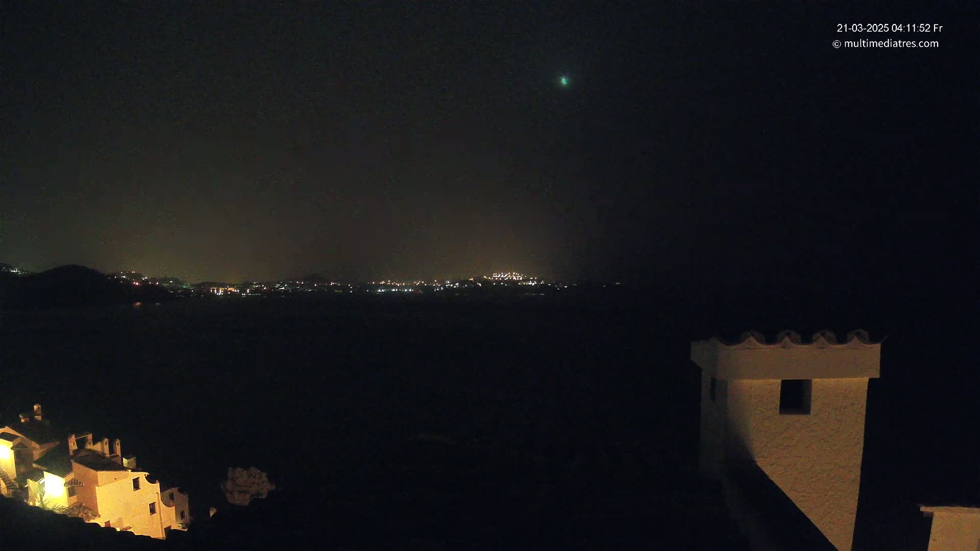 A nighttime long shot shows a distant city skyline illuminated by lights across a dark body of water, with a small, brightly lit building in the foreground under a dark, clear sky featuring a single bright light.