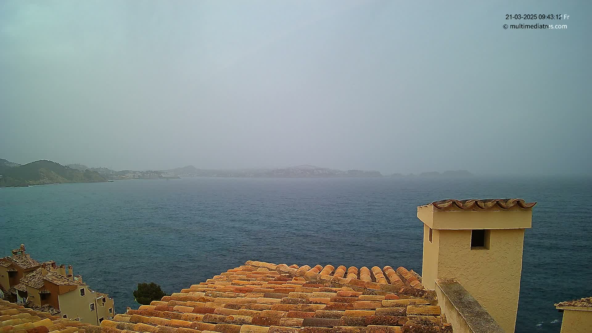 A hazy view of a calm sea stretches to distant, indistinct hills from a vantage point overlooking a terracotta-tiled roof and a short, light-colored chimney.