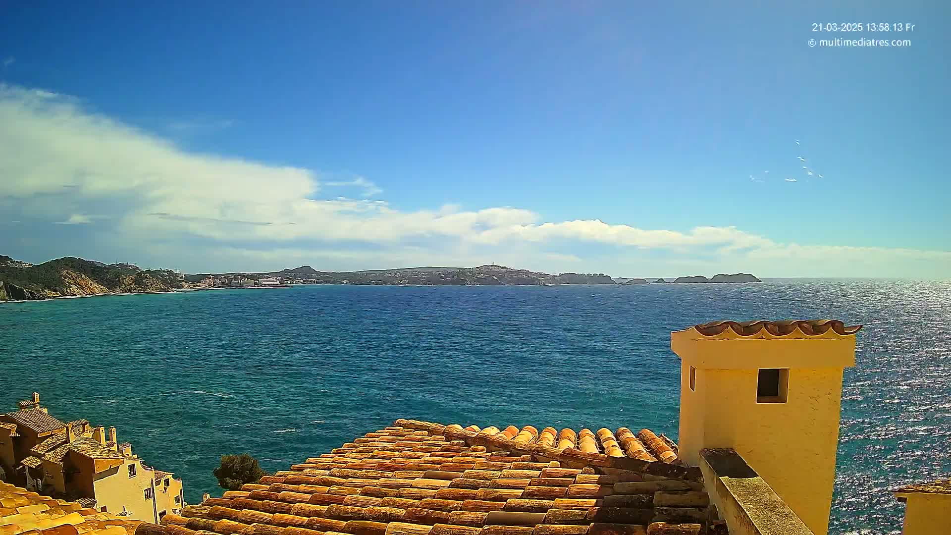 A partly cloudy blue sky overlooks a calm, blue sea from a rooftop with terracotta tiles and a yellow chimney, with distant hills and coastline visible.