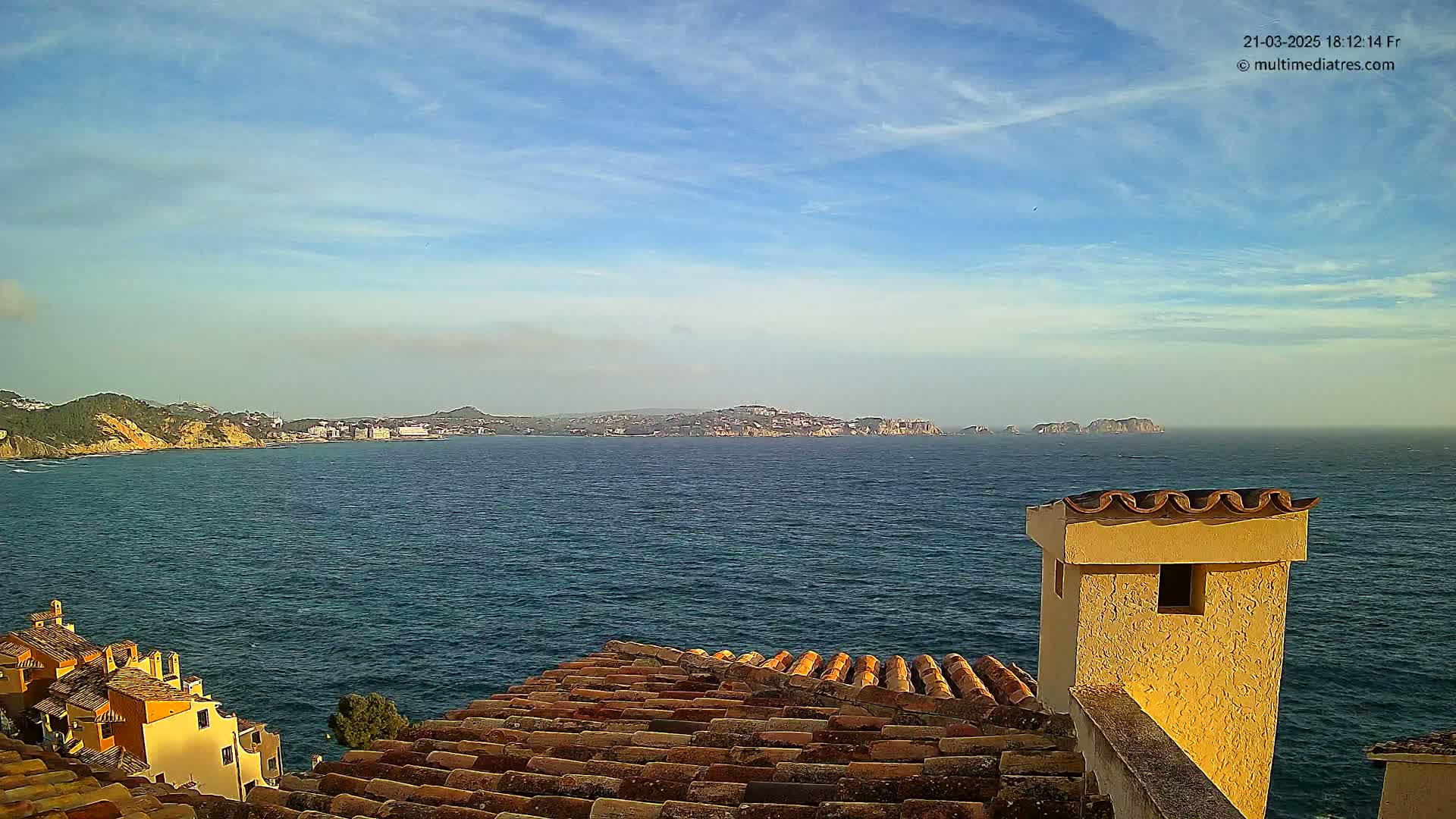 A tiled roof overlooks a calm blue sea and distant coastline under a mostly clear sky.