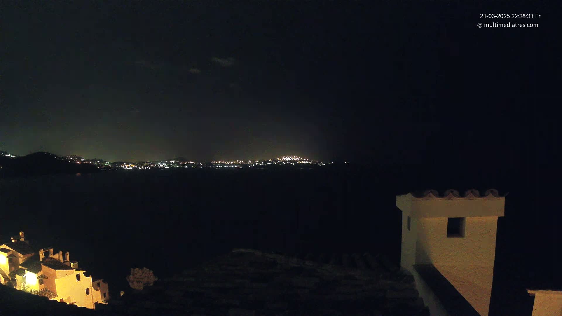 A nighttime view of a city across a dark body of water, partially obscured by darkness, with a portion of a building in the foreground.