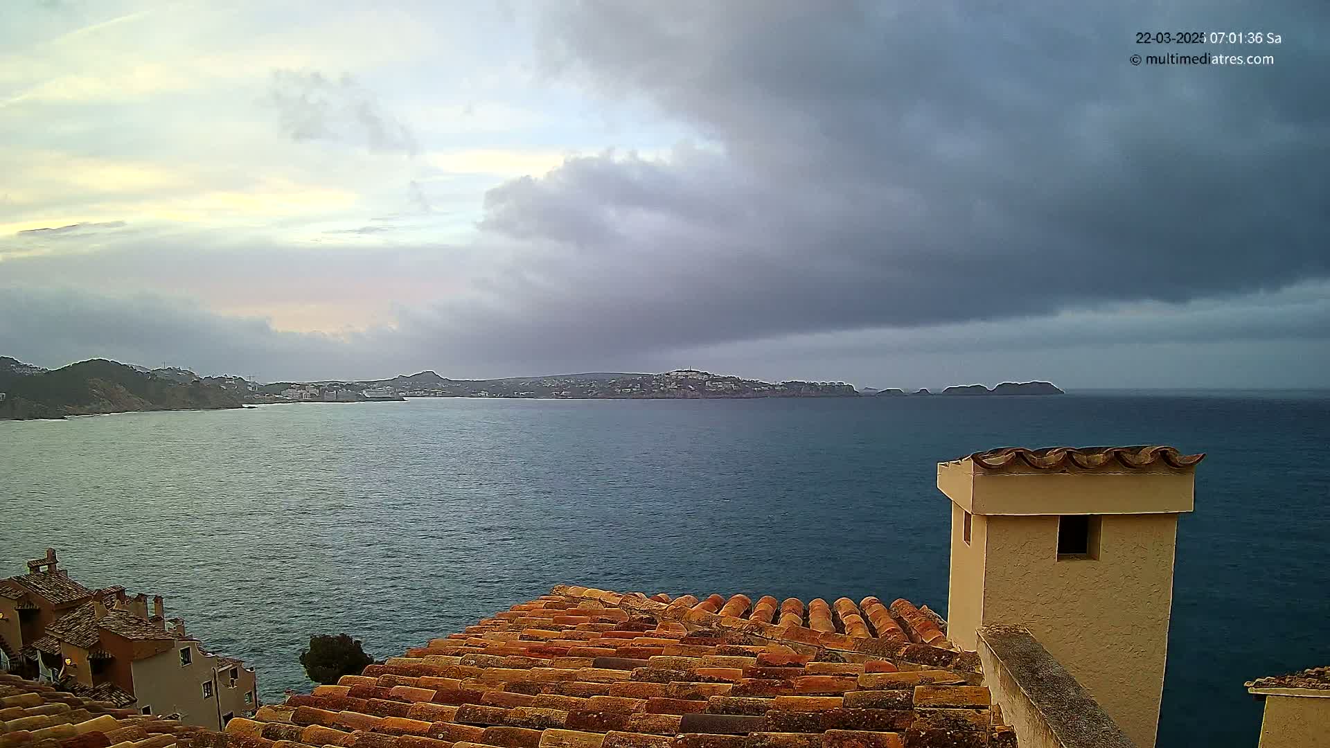 A coastal view shows a calm sea under a partly cloudy sky, with a terracotta roof and chimney in the foreground and a distant town visible on the opposite shore.