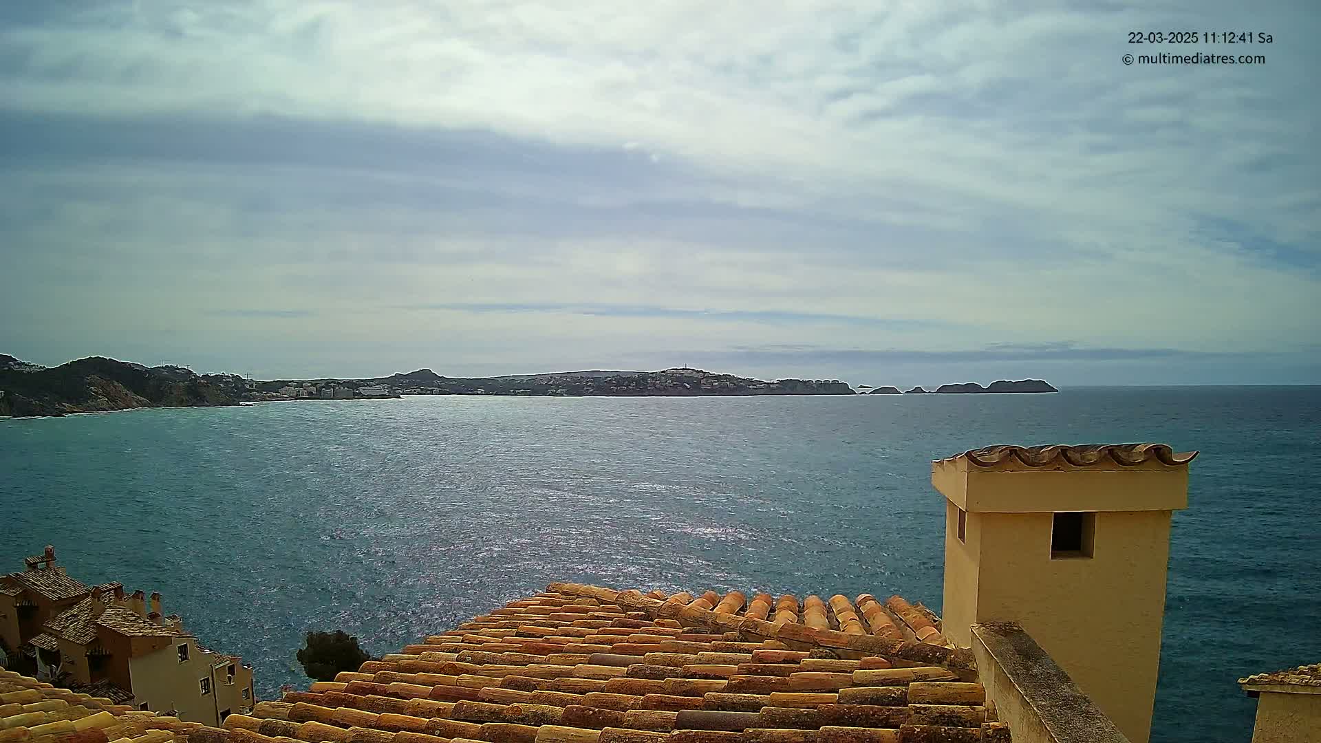 A view from a tiled rooftop shows a calm sea with a distant coastline under a mostly cloudy sky.