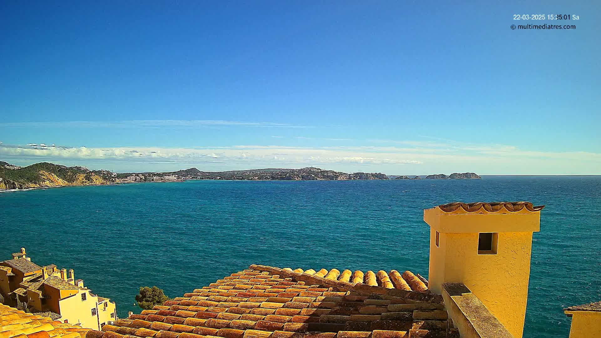 A tiled roof overlooks a calm, turquoise sea under a mostly clear blue sky, with a distant coastline visible on the horizon.