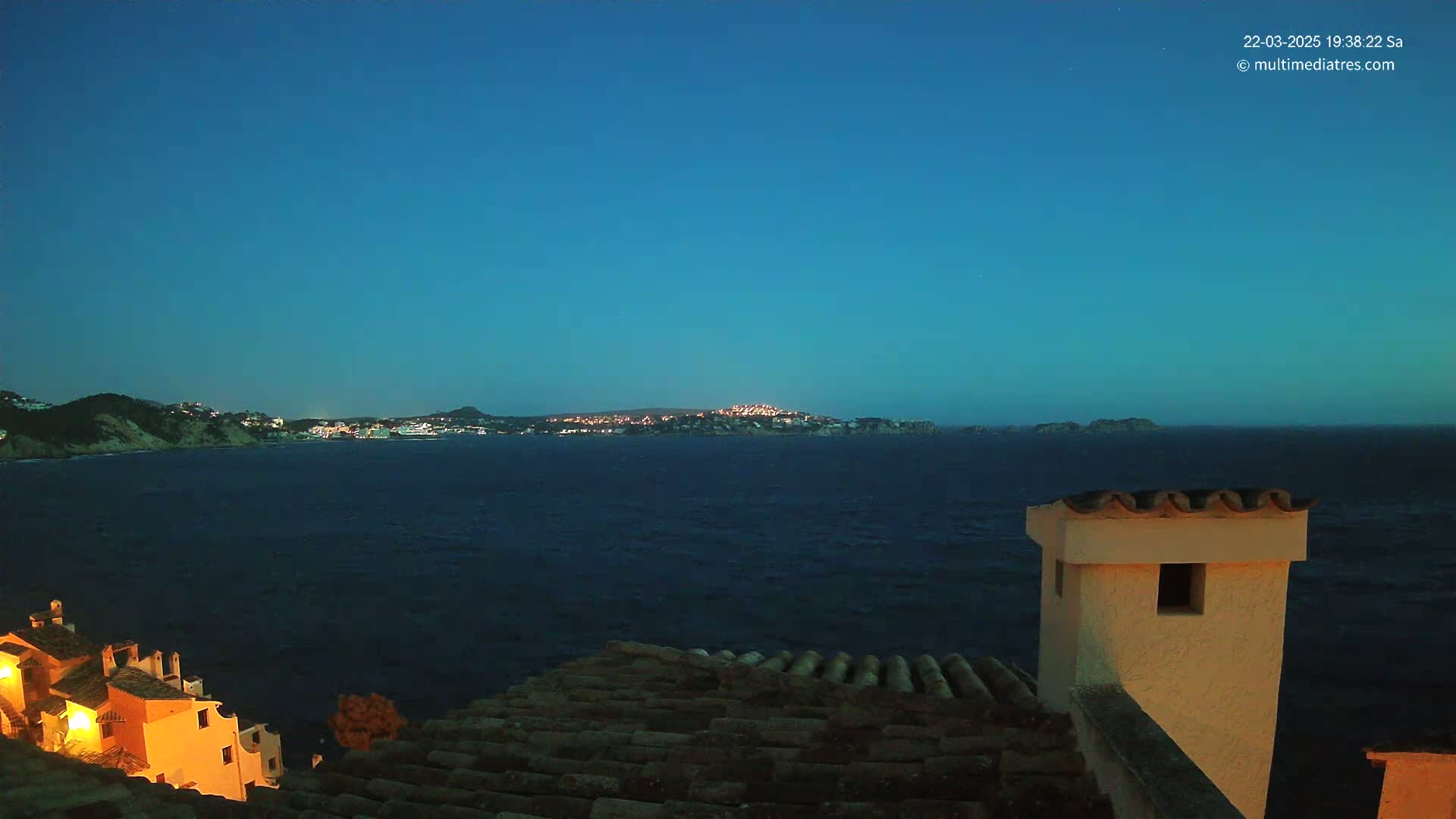 A nighttime coastal view shows a dark blue sea separating a shoreline with illuminated buildings from a distant, similarly lit town under a clear, dark blue sky.