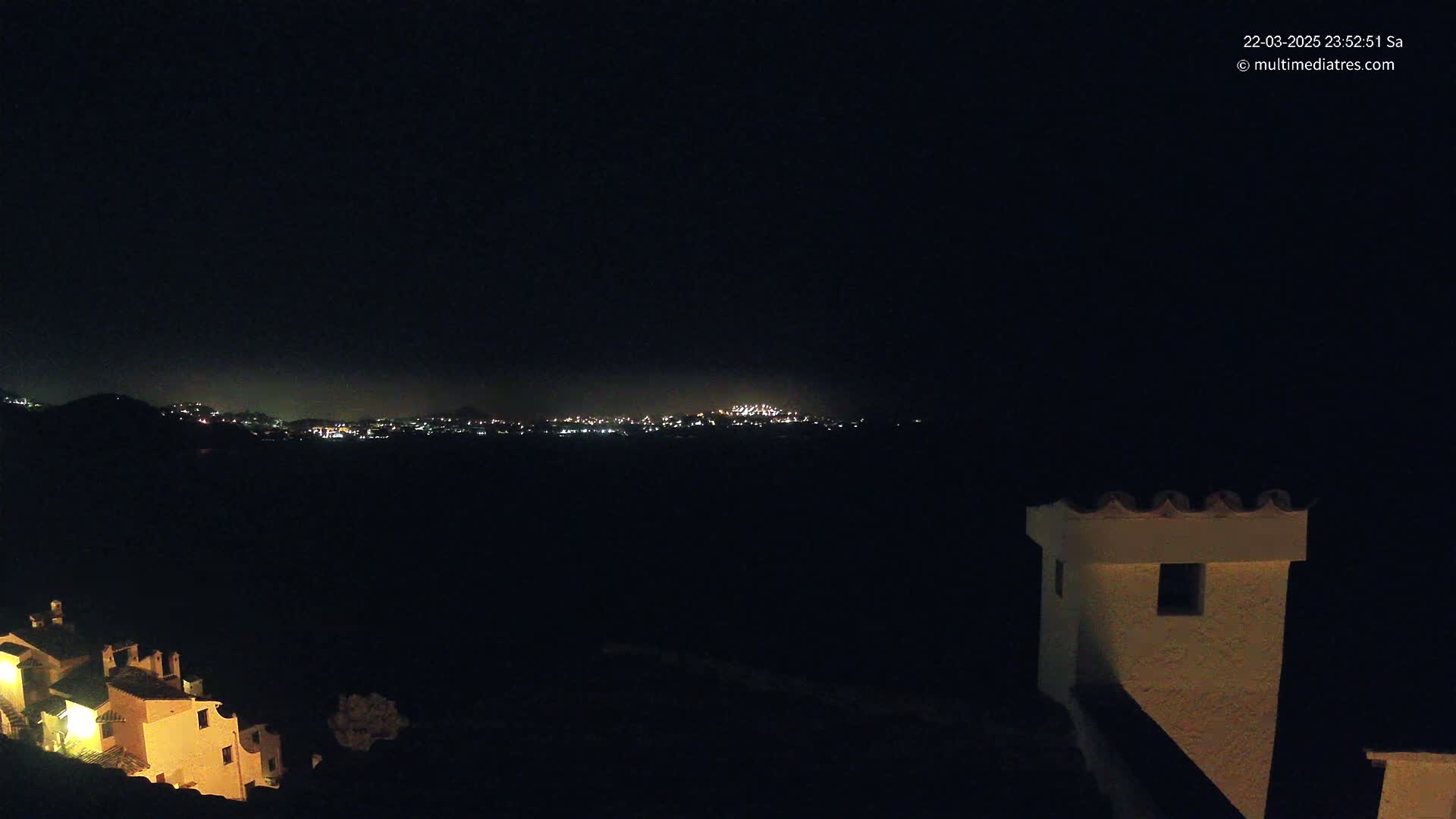 A nighttime view of a distant city skyline illuminated by lights, partially obscured by darkness, with a portion of a building and its illuminated roof visible in the foreground.