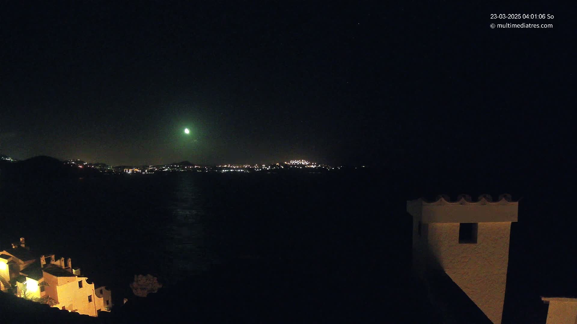 A nighttime coastal scene shows a brightly lit town across a dark body of water, a bright green light in the sky, and a portion of a building in the foreground.