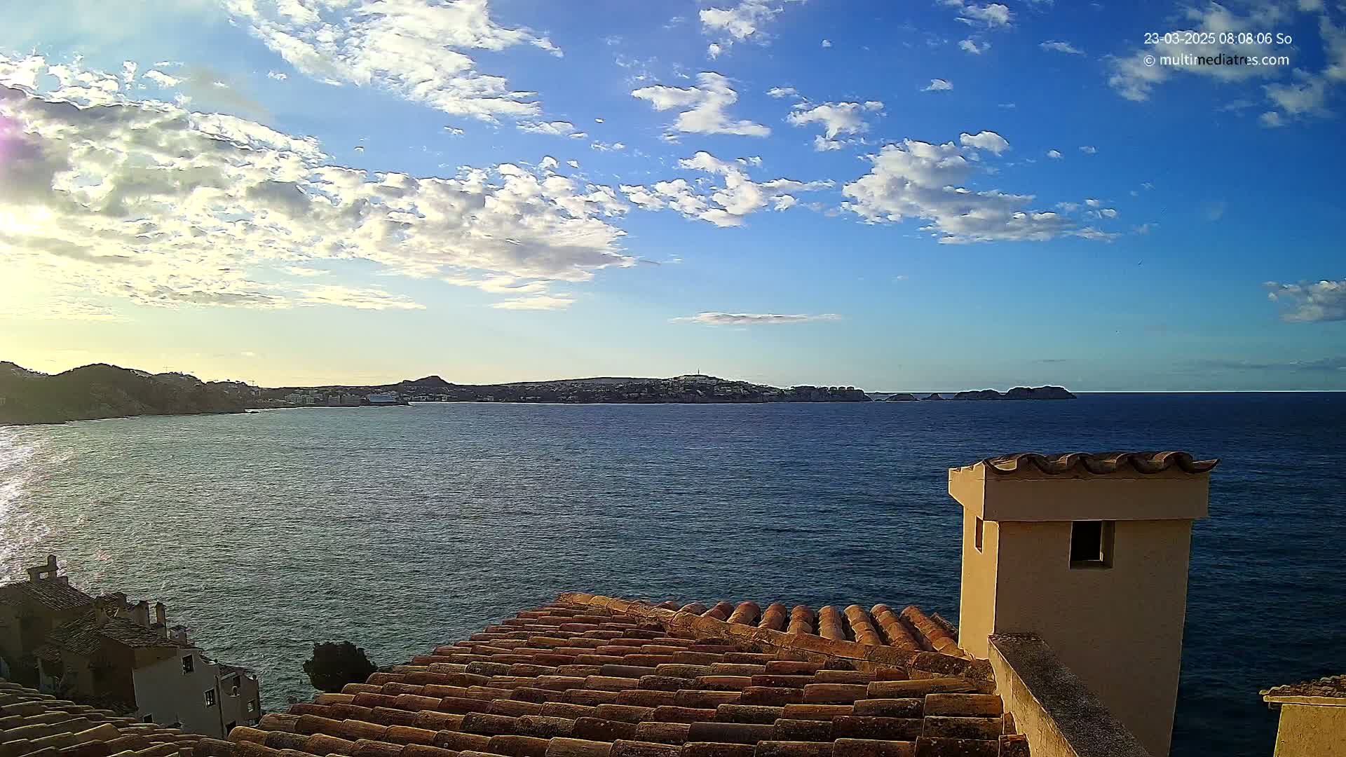 A partly cloudy sky over a calm sea, viewed from a rooftop with terracotta tiles and a small, light-beige chimney.