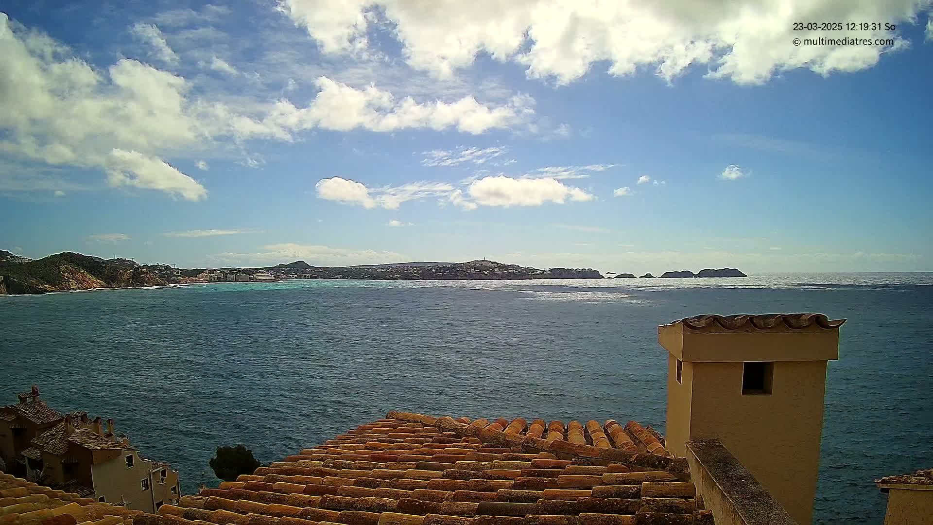 A partly cloudy sky overlooks a calm sea with a distant town and islands visible from a tiled roof with a chimney.