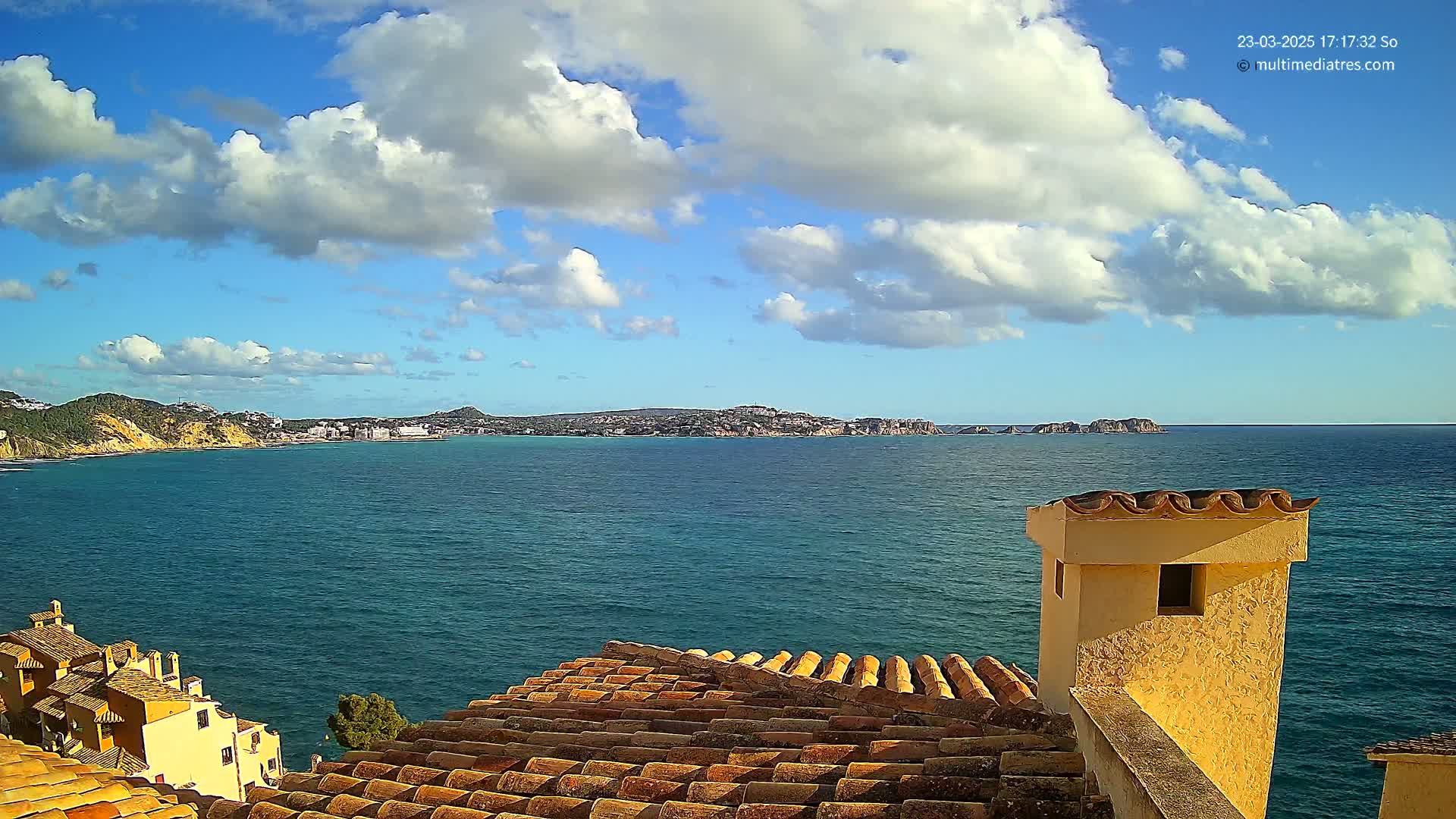 A partly sunny day reveals a coastal scene of teal water, distant landmasses, and a terracotta roof with a yellow chimney, all viewed from a high vantage point.