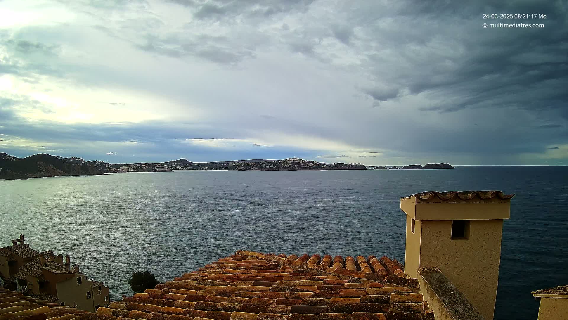 A coastal view shows a calm sea under a mostly cloudy sky, seen from a terracotta roof with a tan chimney.