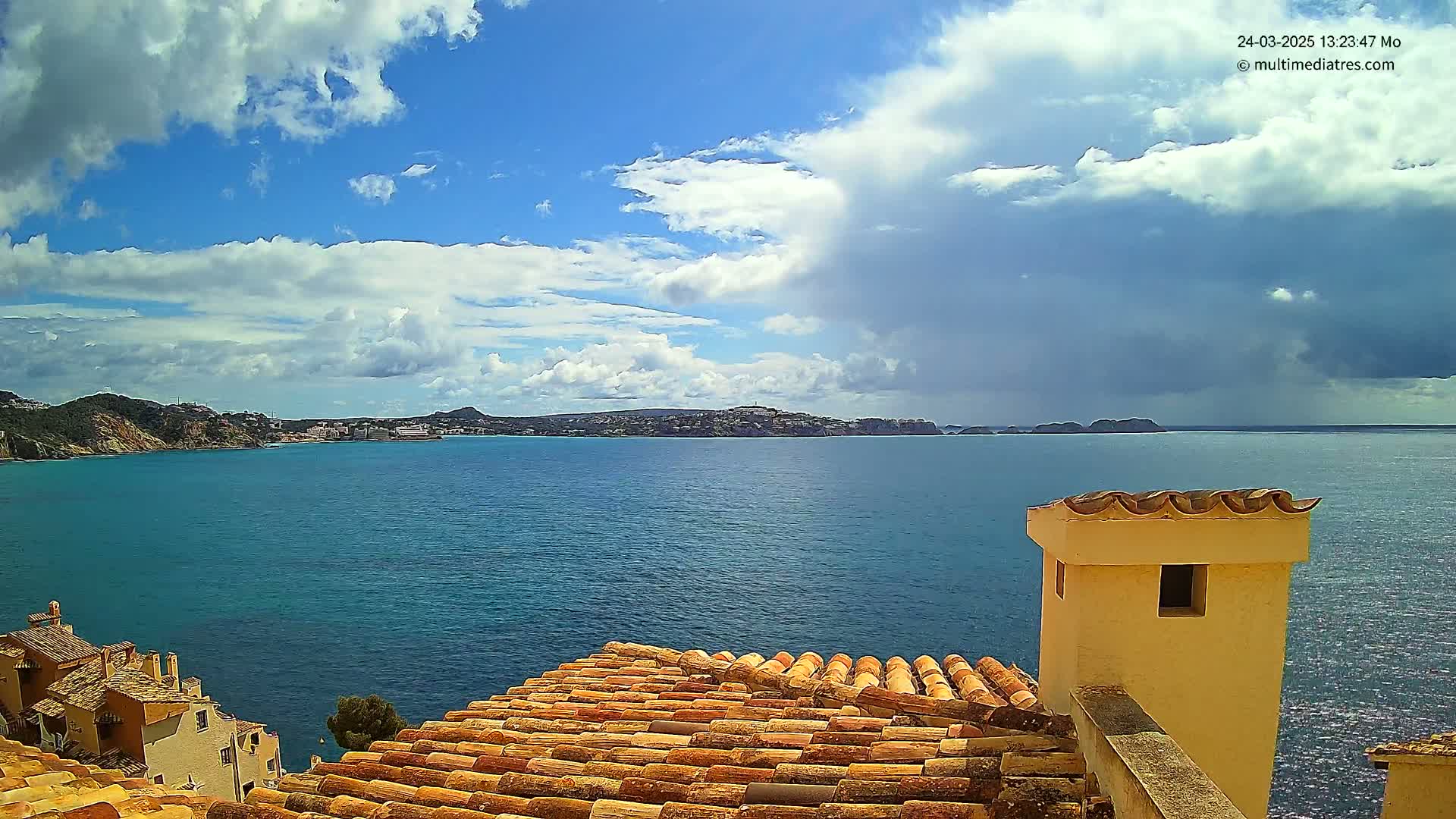 A partly cloudy sky overlooks a calm blue sea, with a coastal town and terracotta rooftops in the foreground.