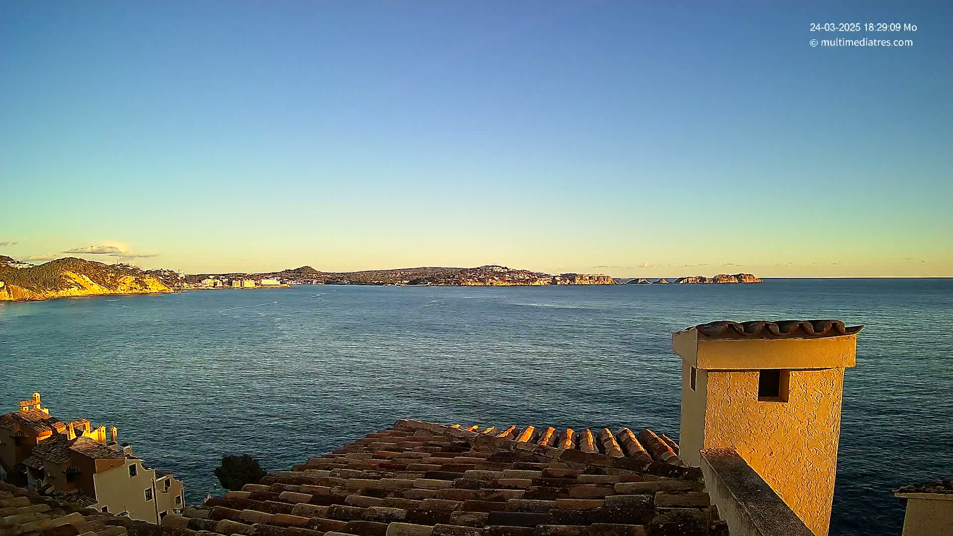 A calm, clear blue sea stretches to a distant coastline under a clear, sunset sky, viewed from a tiled rooftop with a stucco chimney.