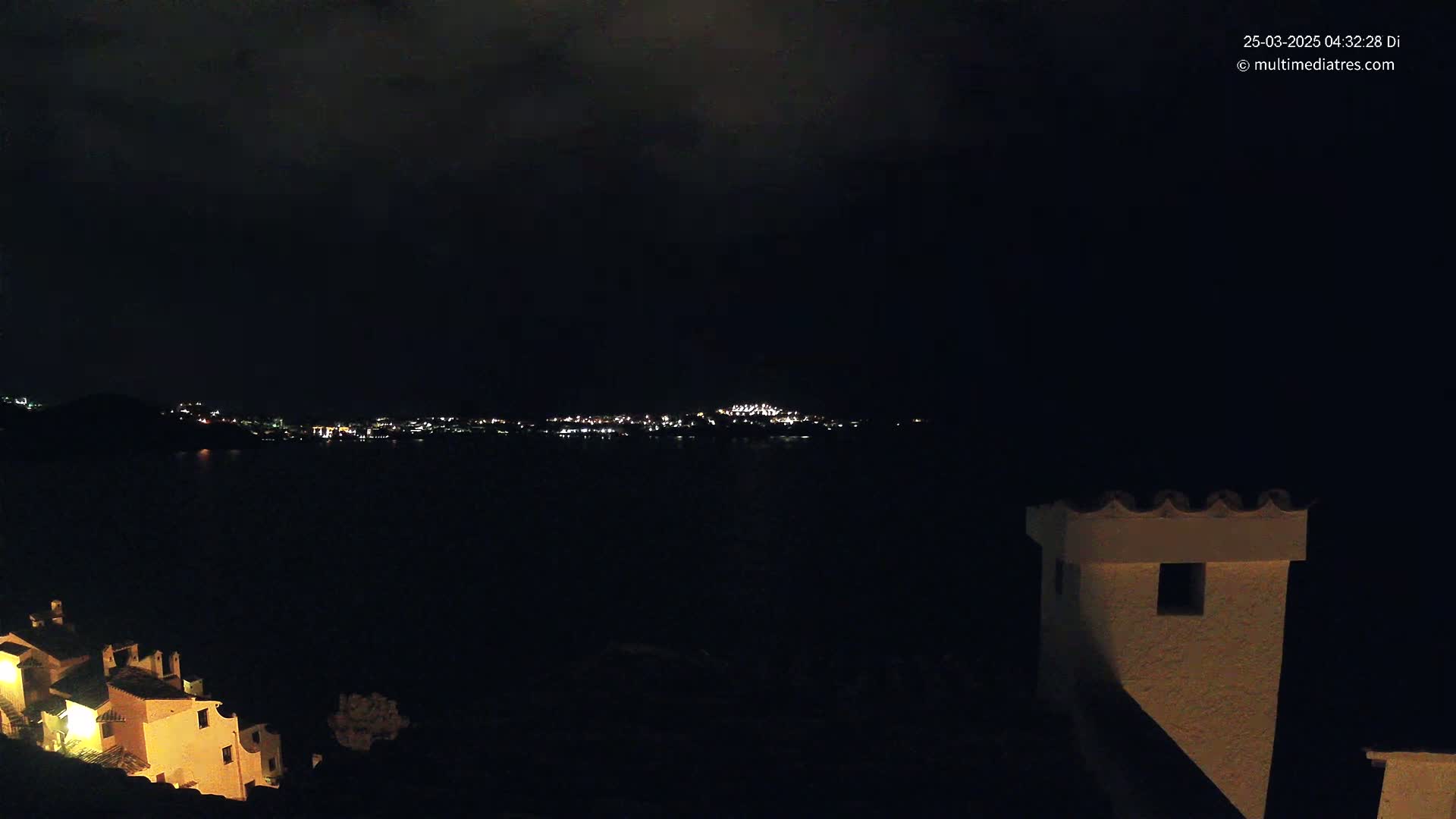 A nighttime view of a city's lights across a dark body of water, with a portion of a building and its illuminated windows in the foreground under a cloudy sky.