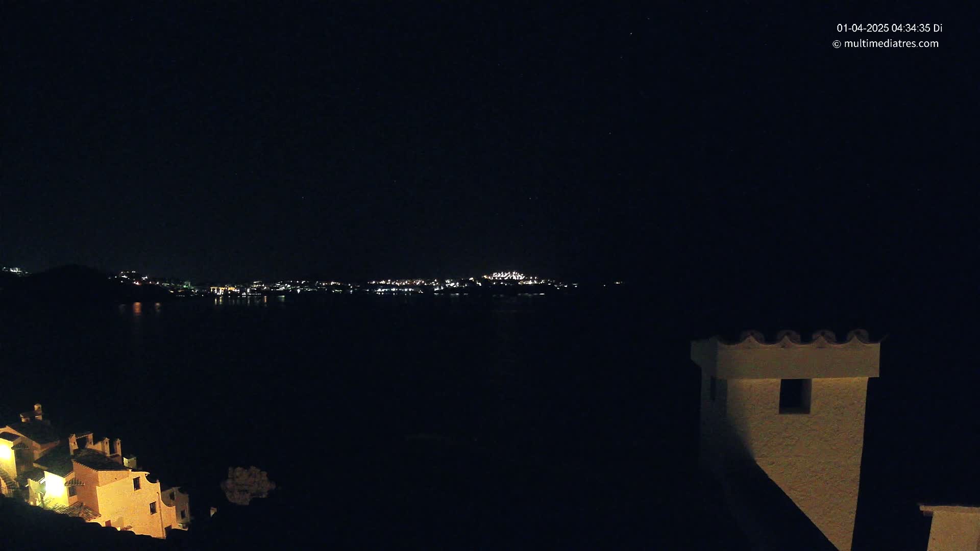 A nighttime view shows a dark body of water separating a dimly lit coastal town from a hilltop with illuminated buildings under a clear, starry sky.