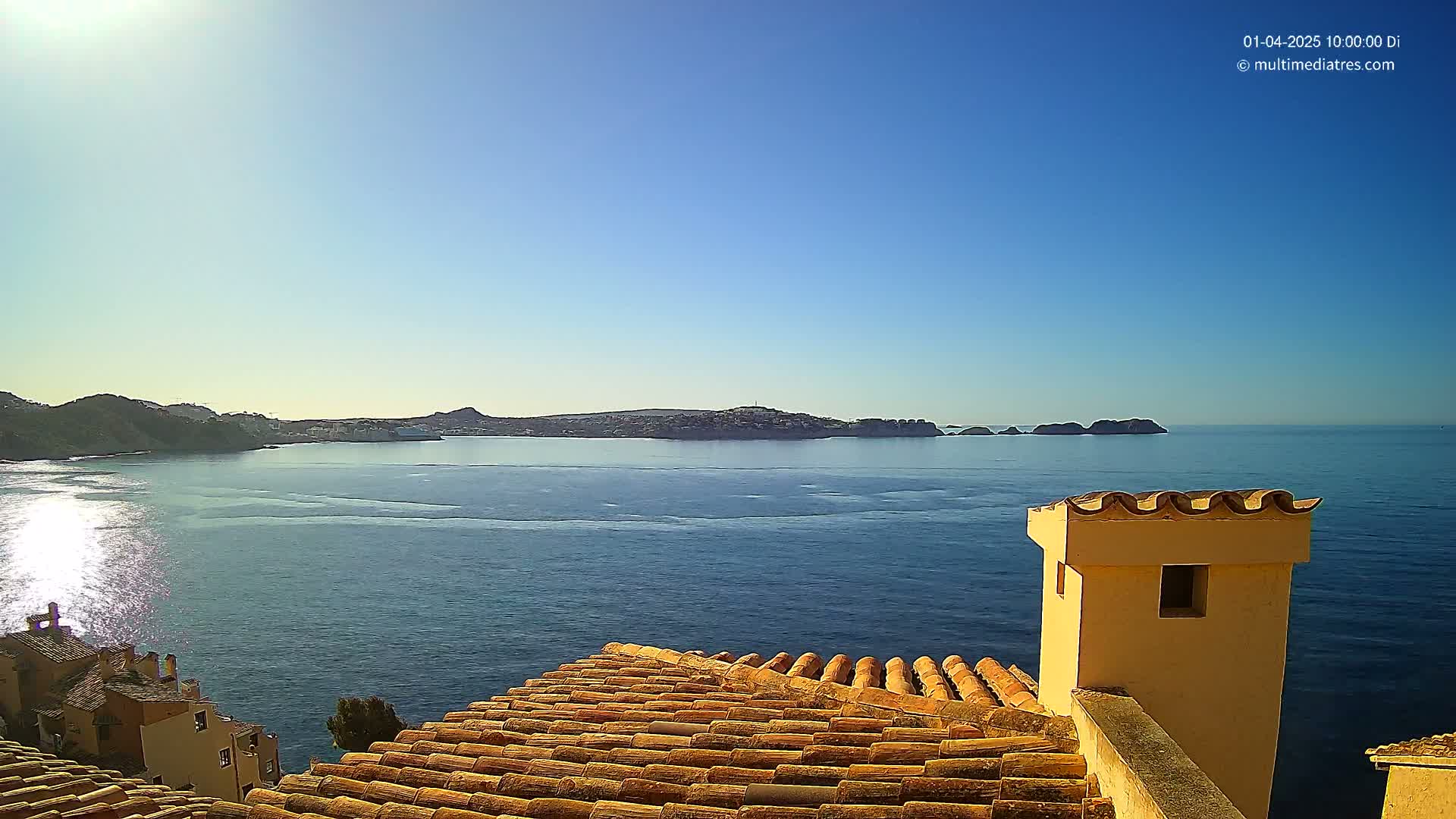 A sunlit coastal scene shows a calm blue sea, a distant island, and a terracotta roof with a chimney in the foreground.