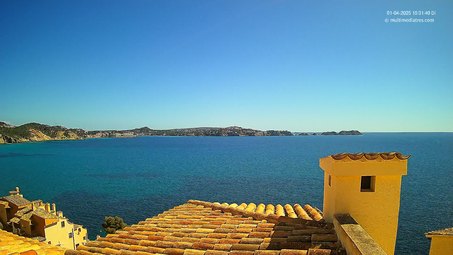 A tiled roof overlooks a calm, blue ocean and distant coastline under a clear blue sky.