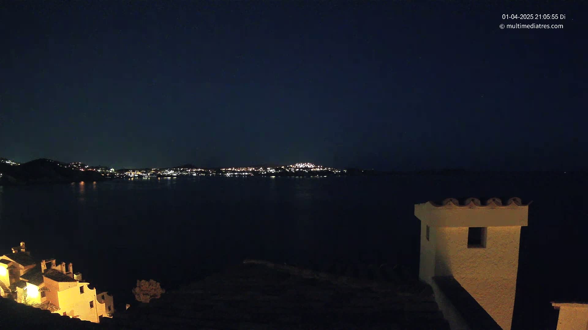 A nighttime view of a dark body of water with a distant, illuminated cityscape and a partially illuminated building in the foreground under a clear, dark sky.