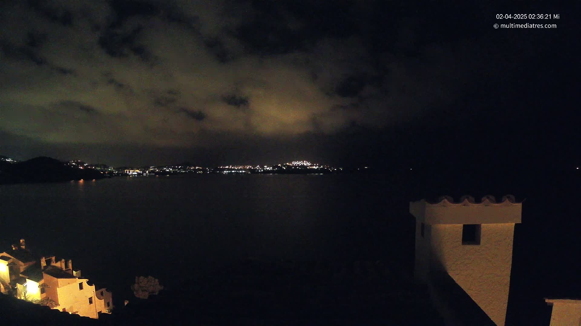 A nighttime view of a dark body of water with a distant city skyline illuminated by lights under a cloudy sky, partially obscuring the stars, and a portion of a building in the foreground.