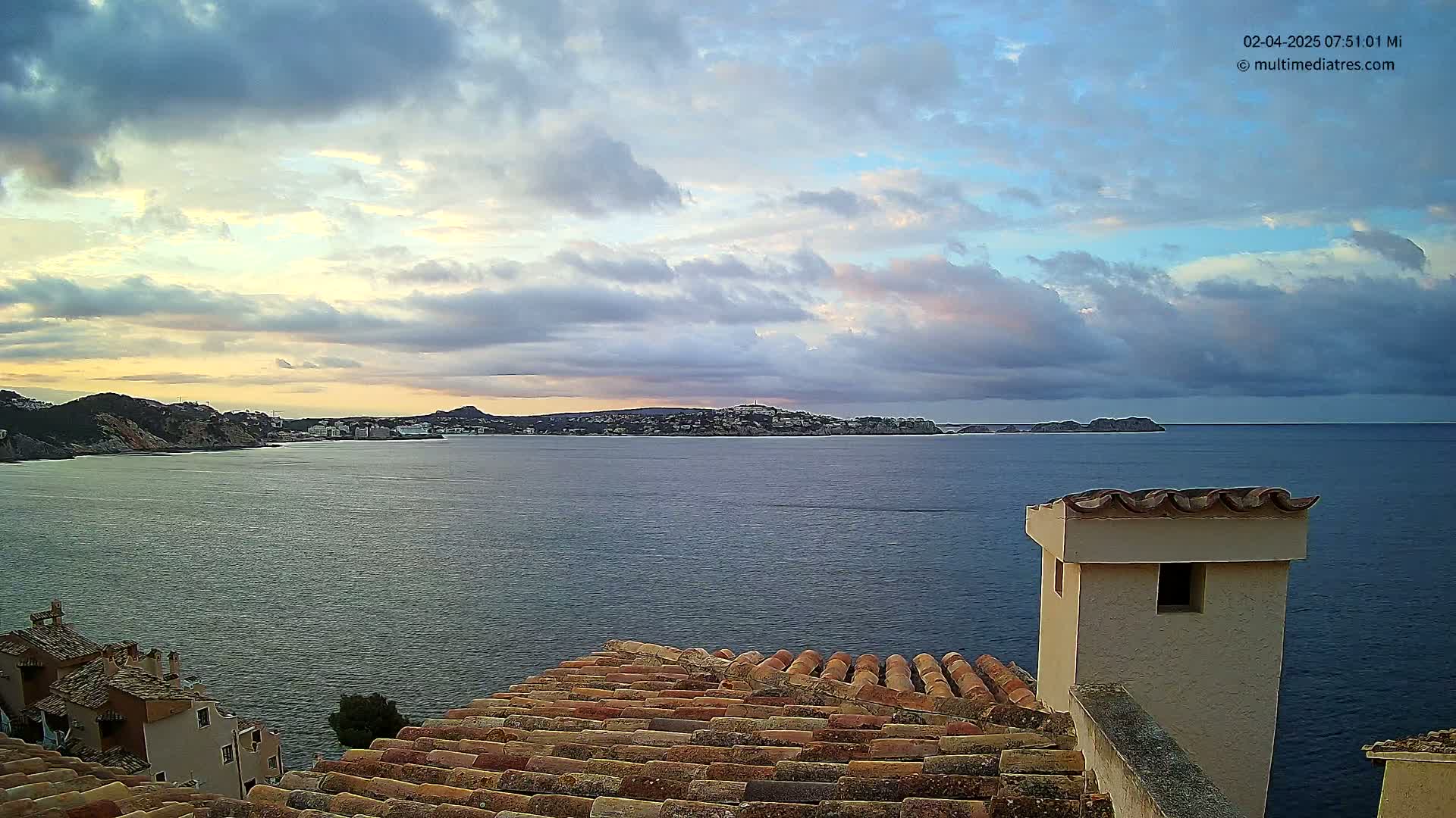 A coastal town is viewed from a rooftop under a partly cloudy sky at sunset, overlooking a calm sea.