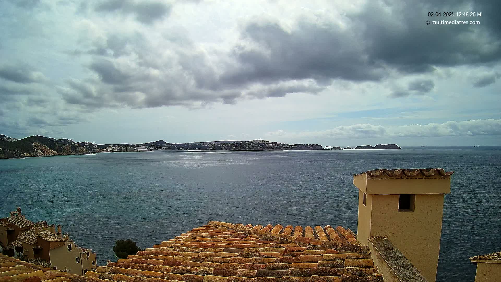 A partly cloudy sky overlooks a calm sea, with a distant coastline and a portion of a tiled roof and chimney in the foreground.