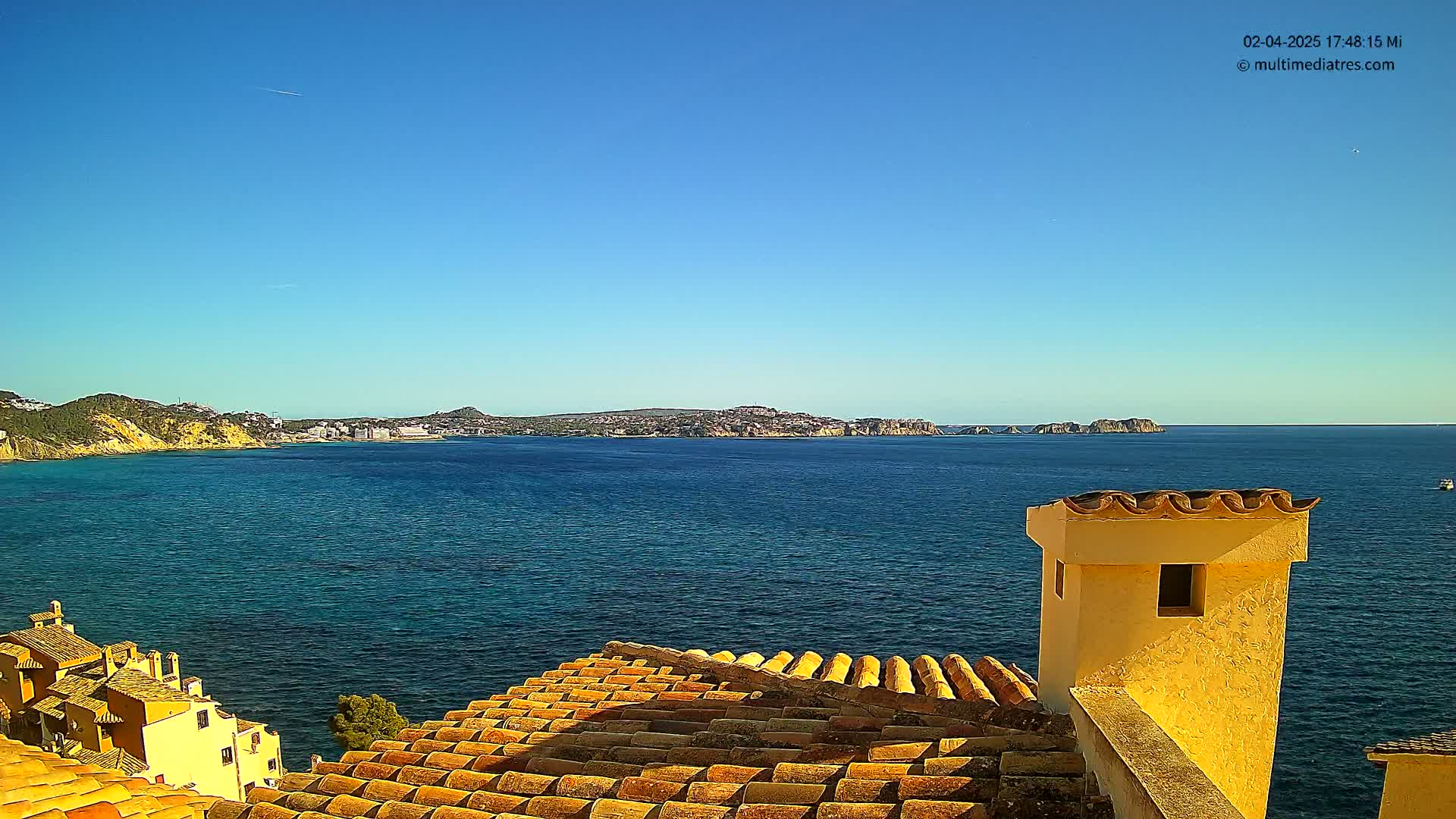 A tiled roof overlooks a calm, blue ocean with a distant coastline under a clear blue sky.