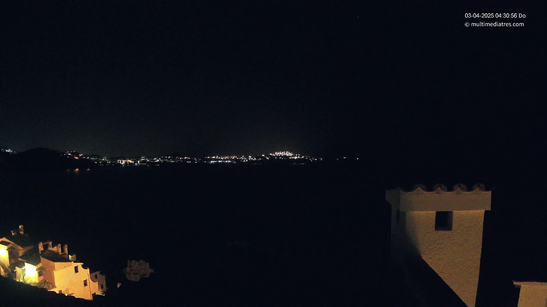 A nighttime view of a distant city skyline illuminated by lights across a dark body of water, partially obscured by darkness, with a portion of a lit building and chimney visible in the foreground under a clear, dark sky.