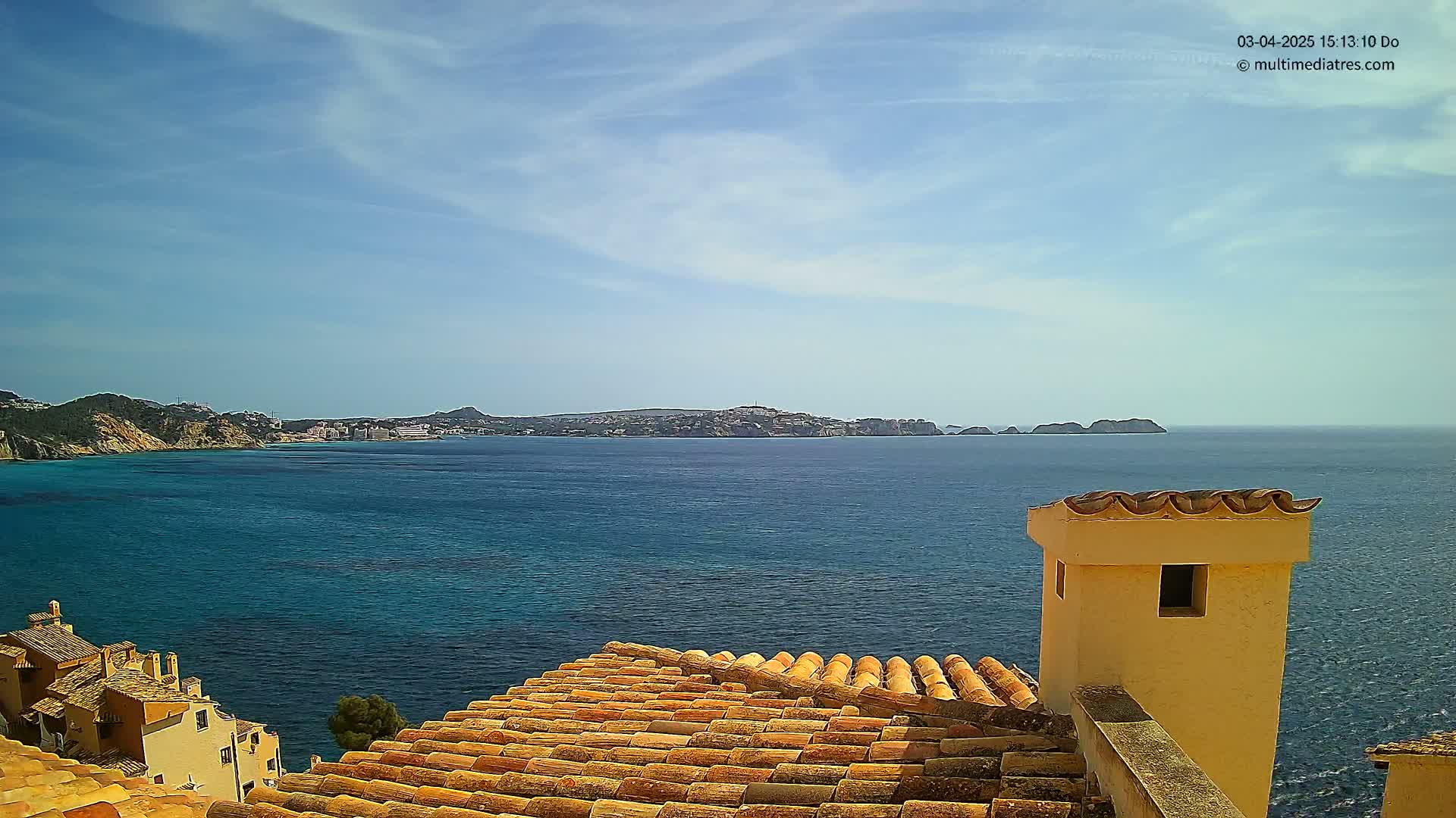 A terracotta roof overlooks a calm, blue sea and distant coastline under a mostly sunny sky.